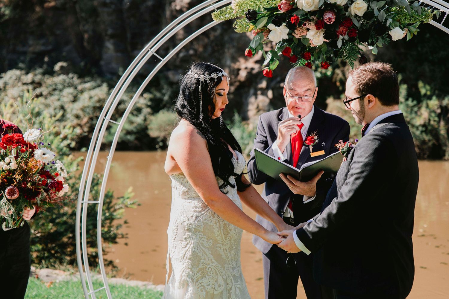 A couple exchanges wedding vows under a white floral arch during an outdoor garden ceremony.