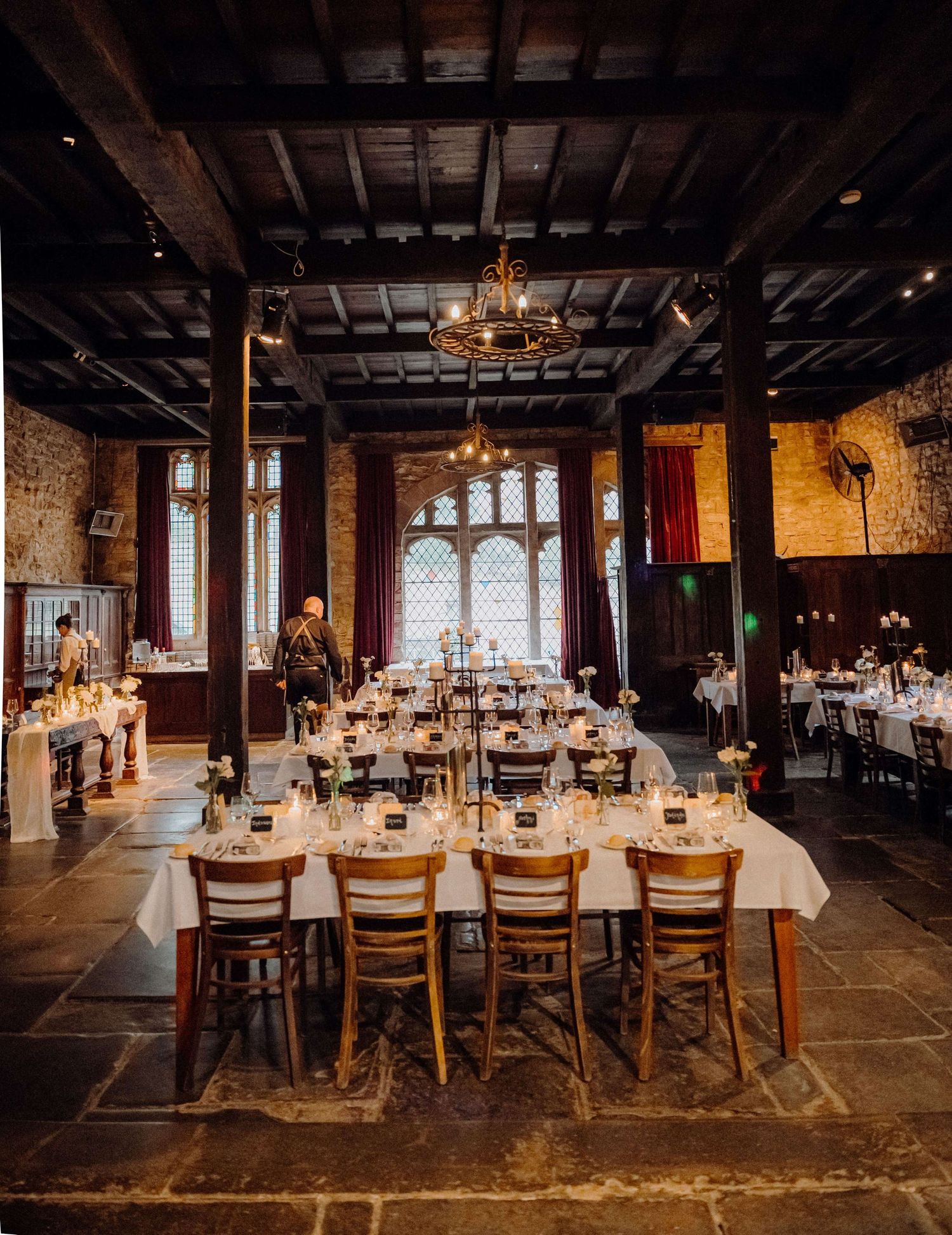Long rustic wooden banquet tables arranged in rows inside a dark historic venue with elegant window lighting and place settings.