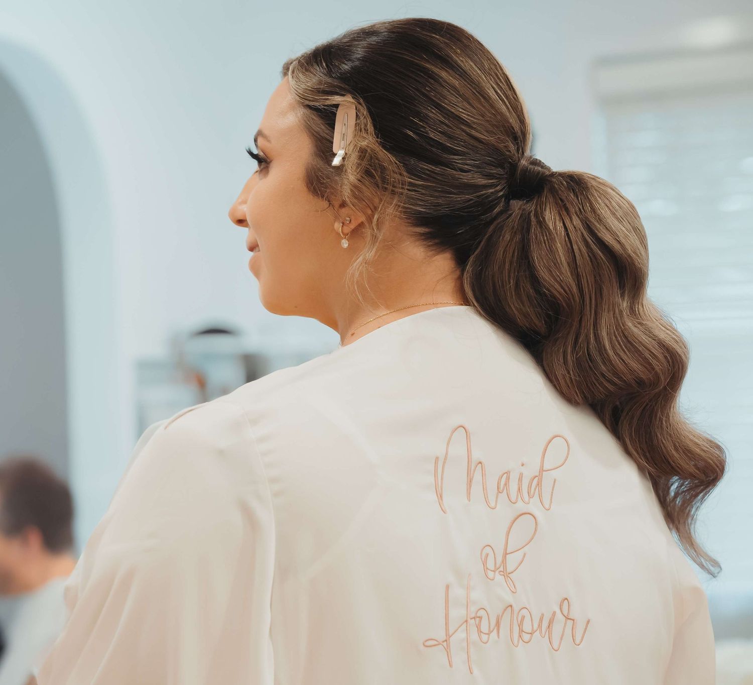 A ponytailed hairstyle from behind with 'Maid of honour' text written in rose gold on a white satin robe.