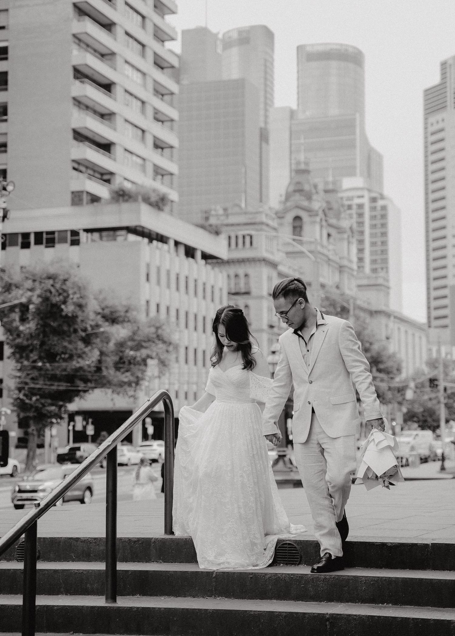 A couple in wedding attire descends stone steps in a cityscape with modern buildings and a church in the background.