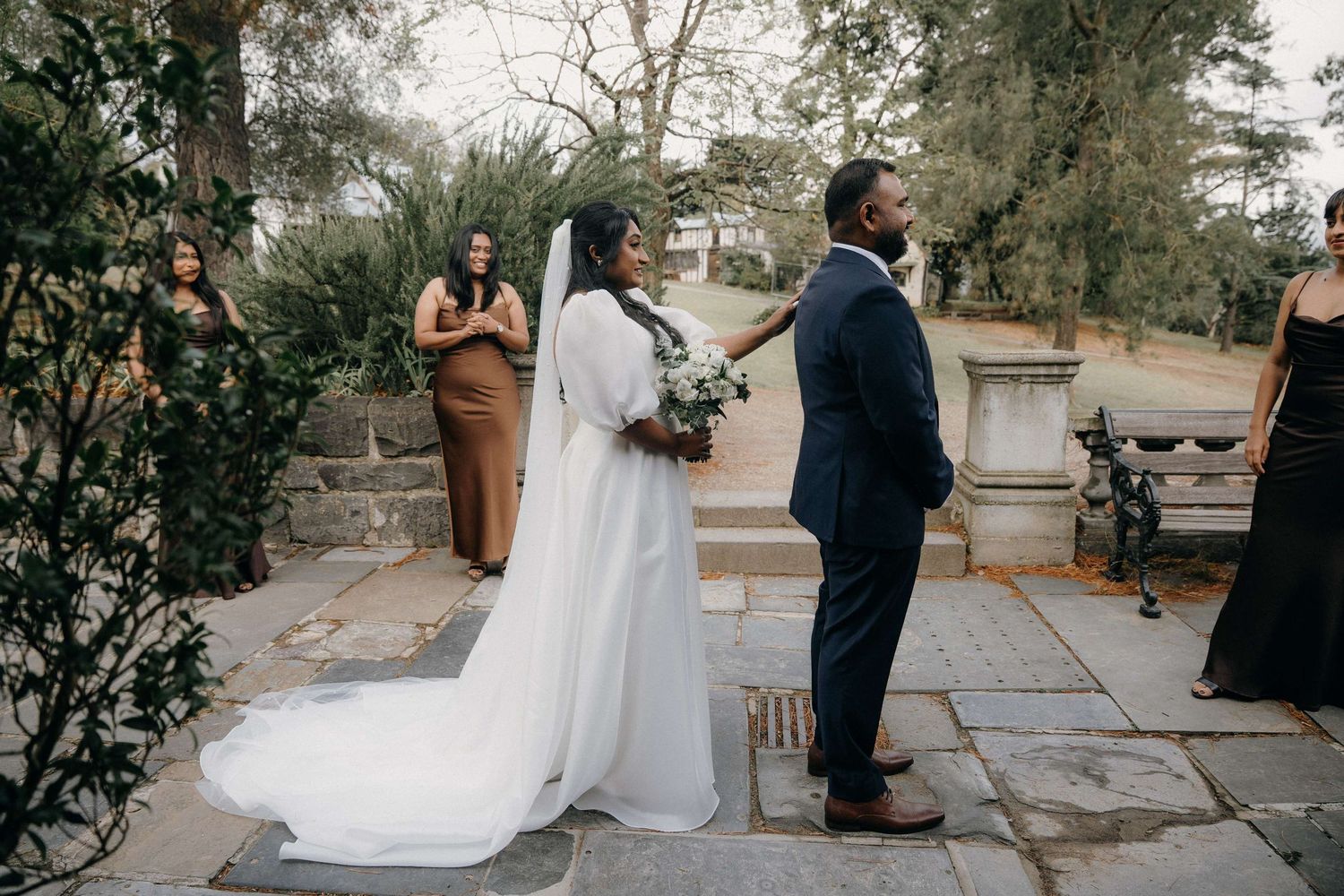 Wedding ceremony moment outdoors with couple and attendants on stone terrace surrounded by greenery.