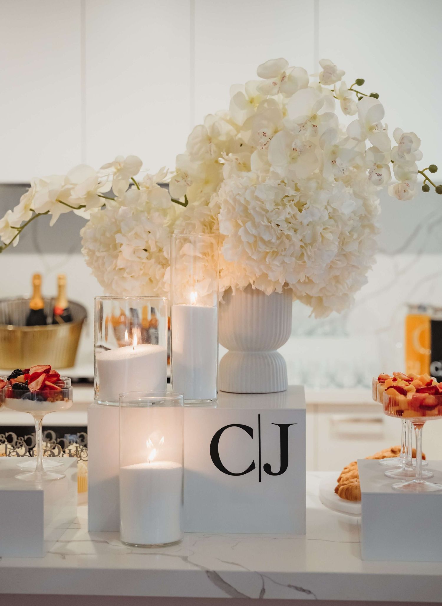 Elegant white hydrangea floral centerpiece with glowing candles and monogram display on a modern white table setting.