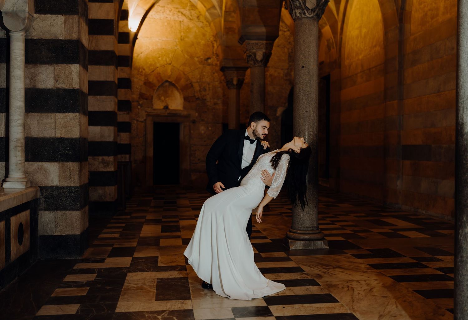 A couple in formal wedding attire dances elegantly in a historic stone hallway with dramatic lighting and striped columns.