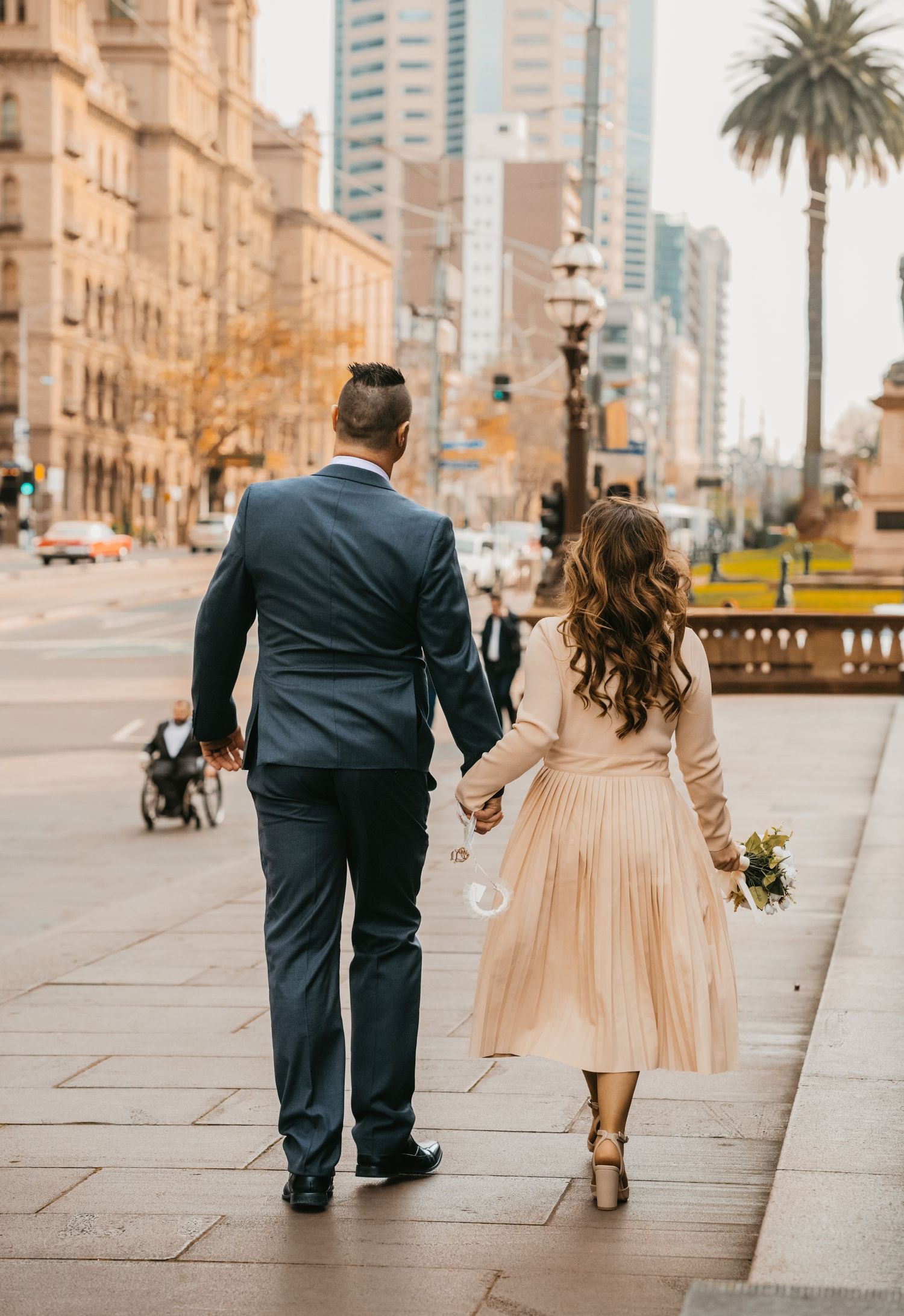 A couple in formal attire walks along an urban sidewalk with palm trees and buildings in the background.