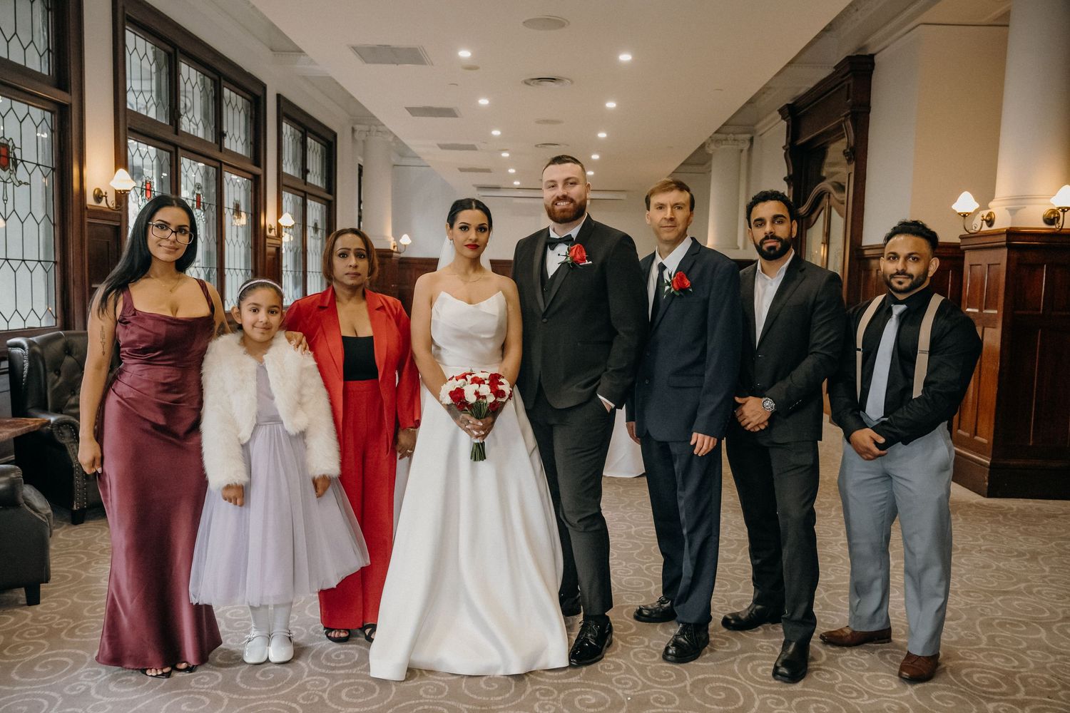 Wedding party poses indoors, bride in white gown with bouquet, groom in suit, bridesmaids in red, groomsmen in dark suits.
