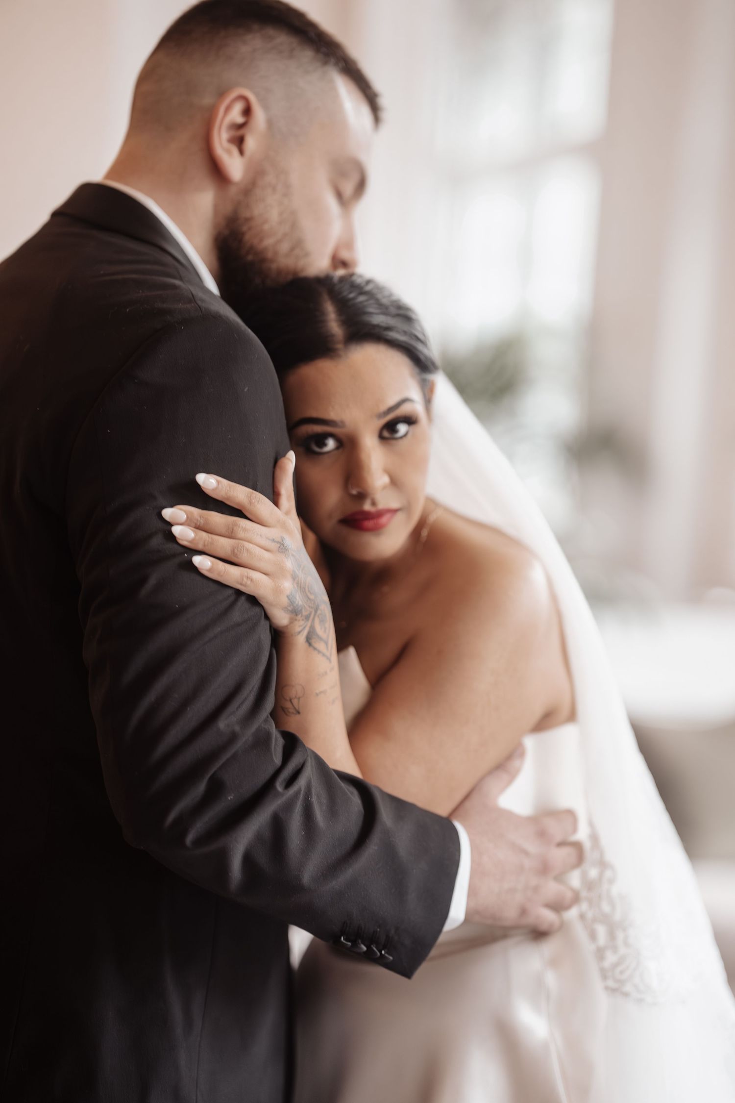 Bride in white gown and veil embraces groom in black suit during romantic wedding portrait indoors.