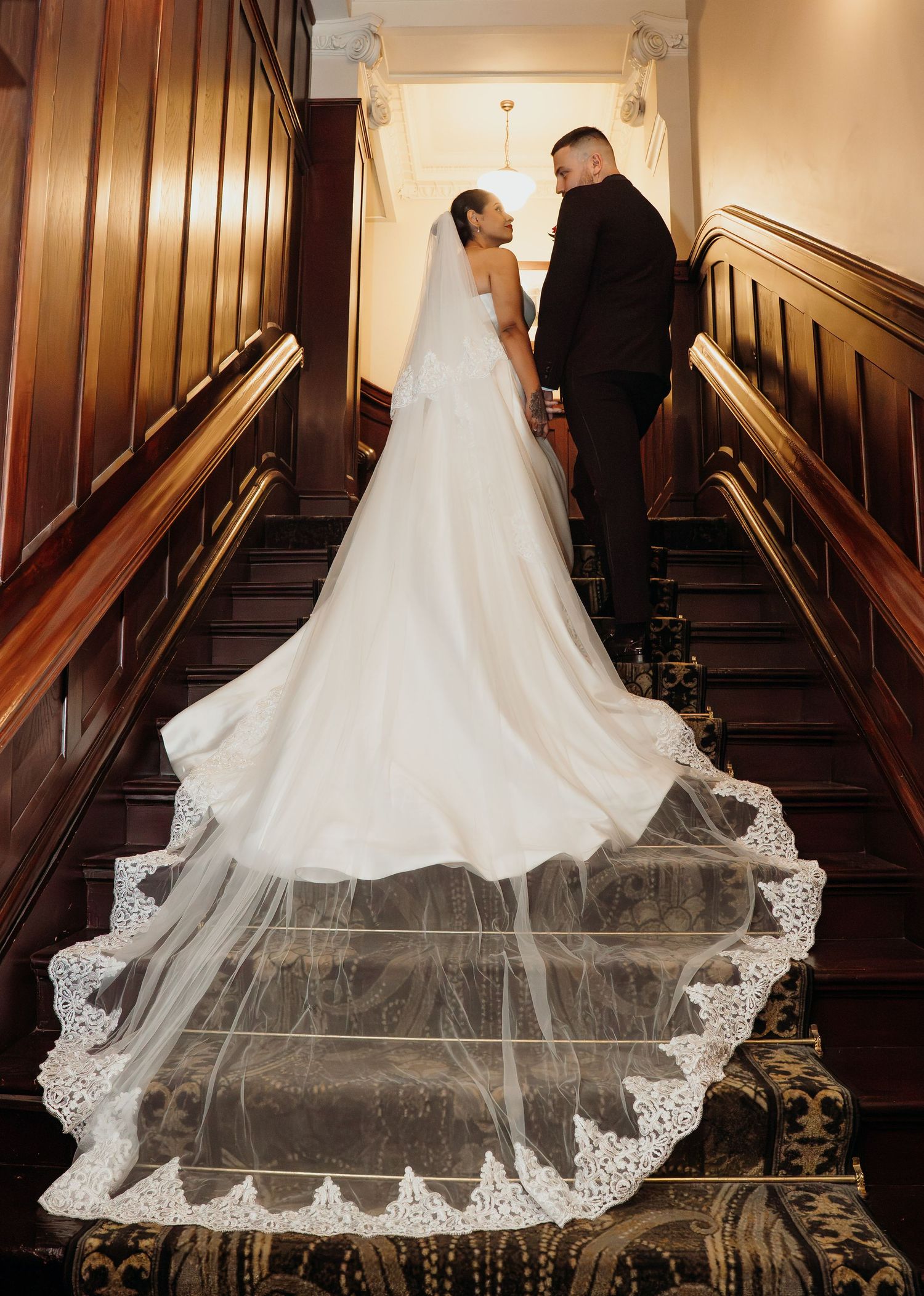 Bride in white gown with lace-trimmed cathedral veil stands with groom on elegant wooden staircase.