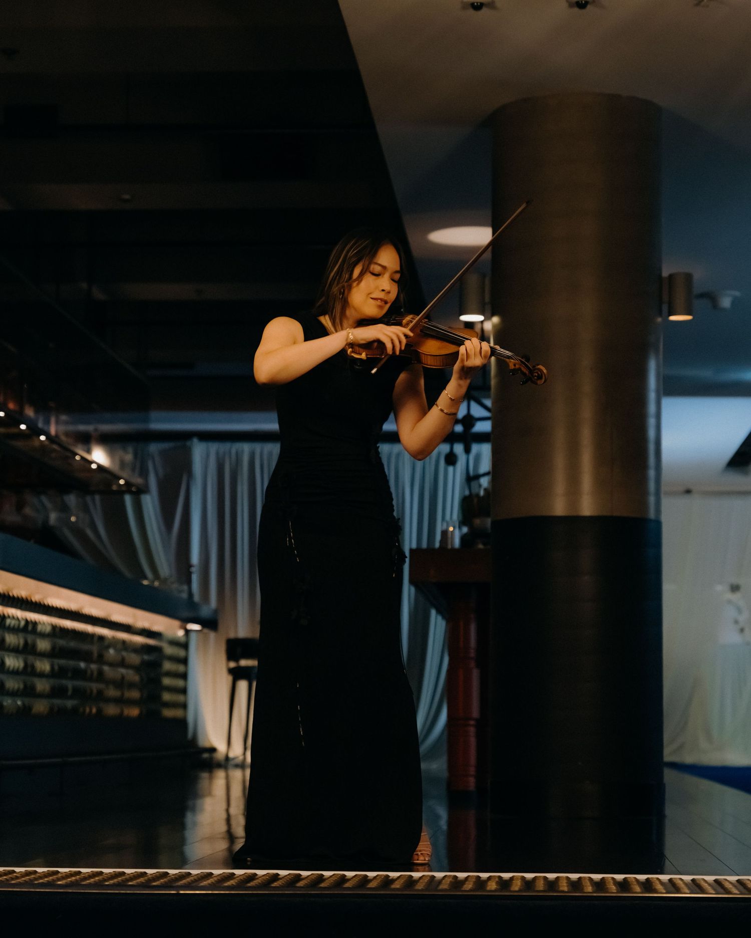 Violinist in black dress performs on stage in dramatic lighting against dark background.