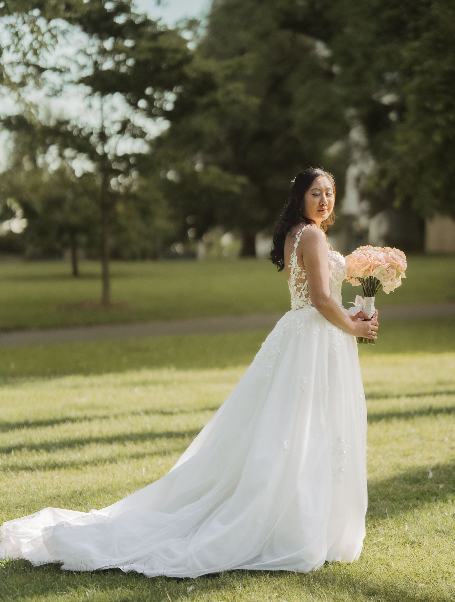 A bride in a flowing white wedding gown poses gracefully on a sunlit grassy lawn during a beautiful outdoor photo session.