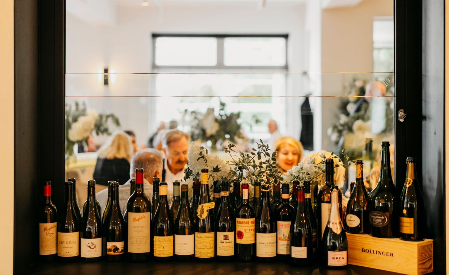 A long row of wine bottles displayed on a wooden shelf against a white wall in a restaurant or wine bar setting.