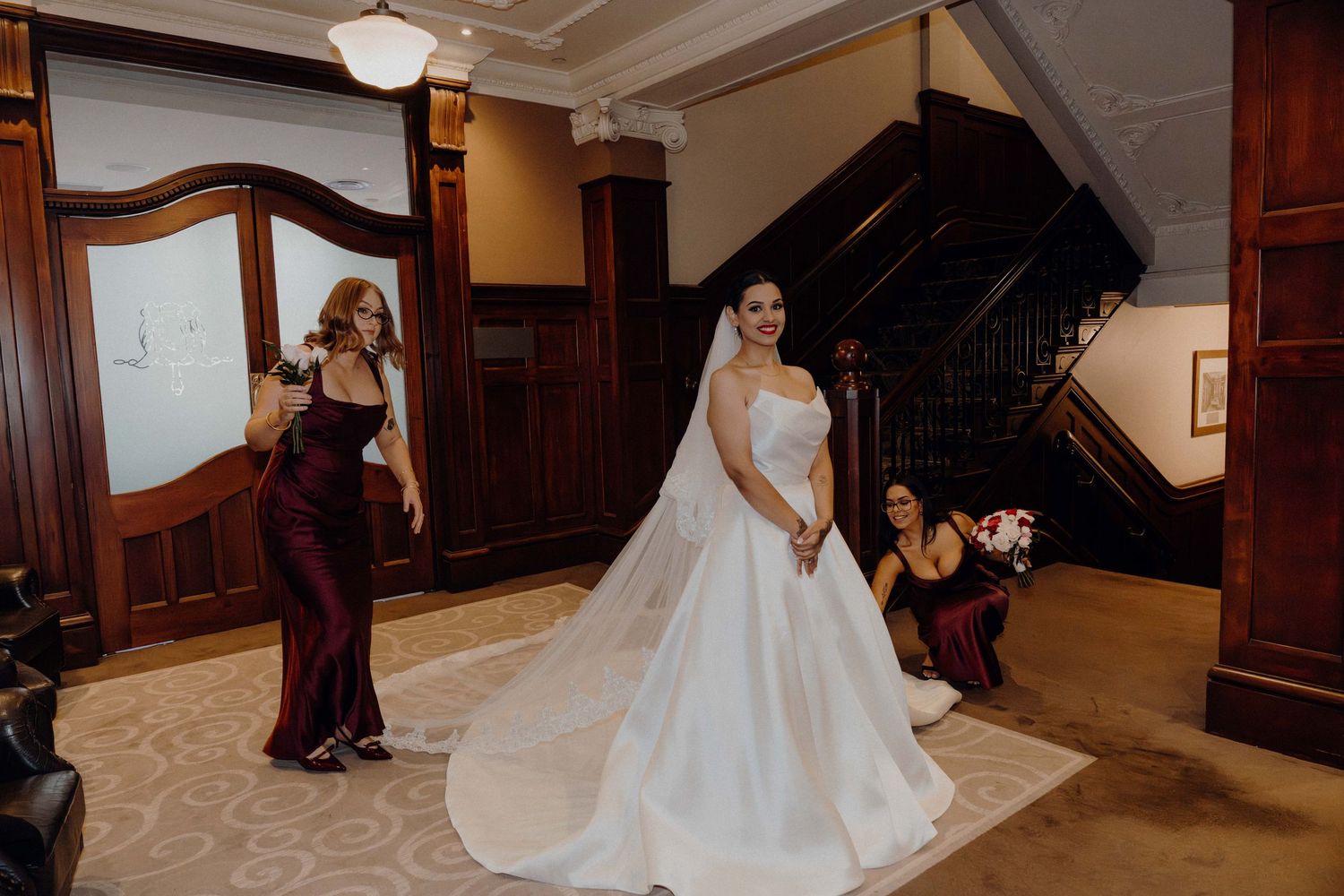 Bride in elegant white ball gown with veil stands in ornate venue lobby while bridesmaids in burgundy dresses assist her.