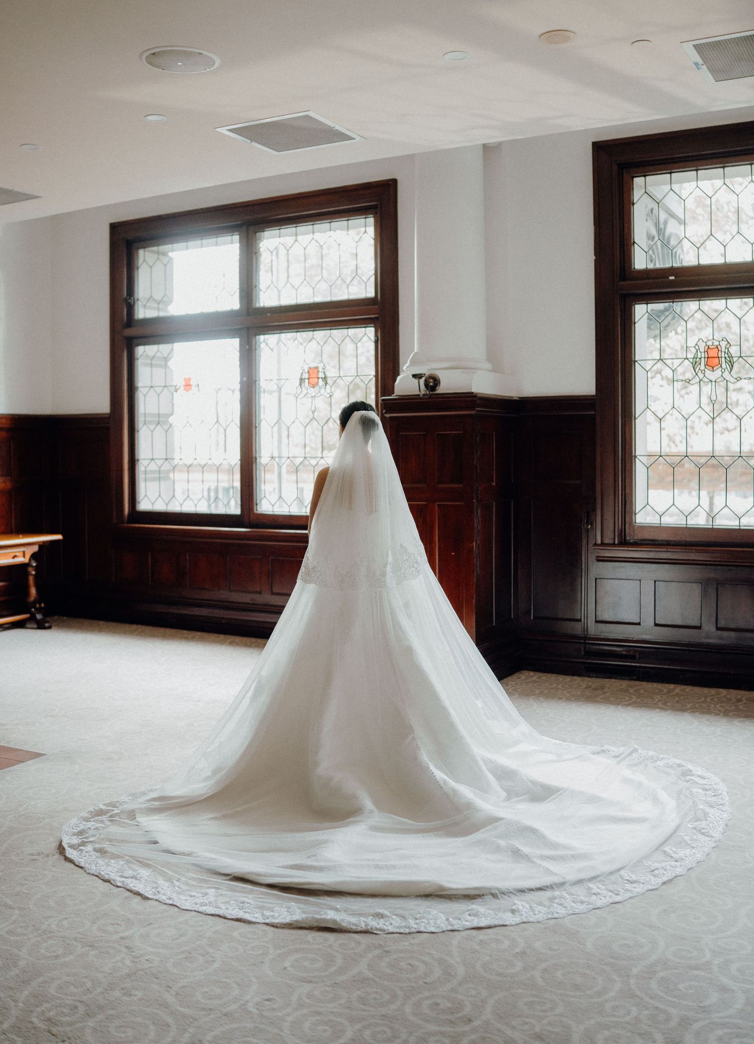 Bride in a white gown with a long lace-trimmed cathedral veil stands before stained glass windows in an elegant room.