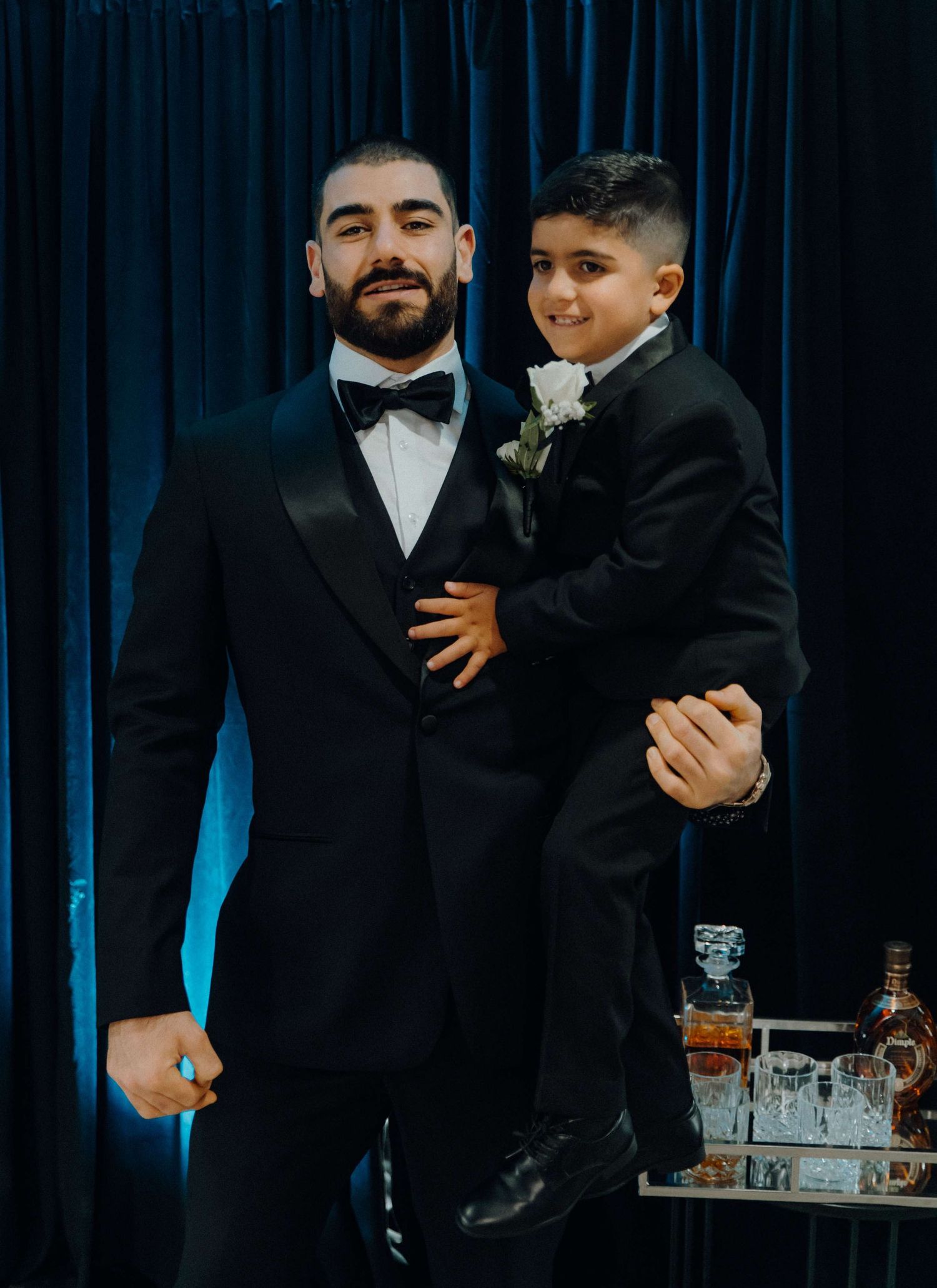 A pair in matching black tuxedos and bowties pose together by a dark blue curtain near a drink display.
