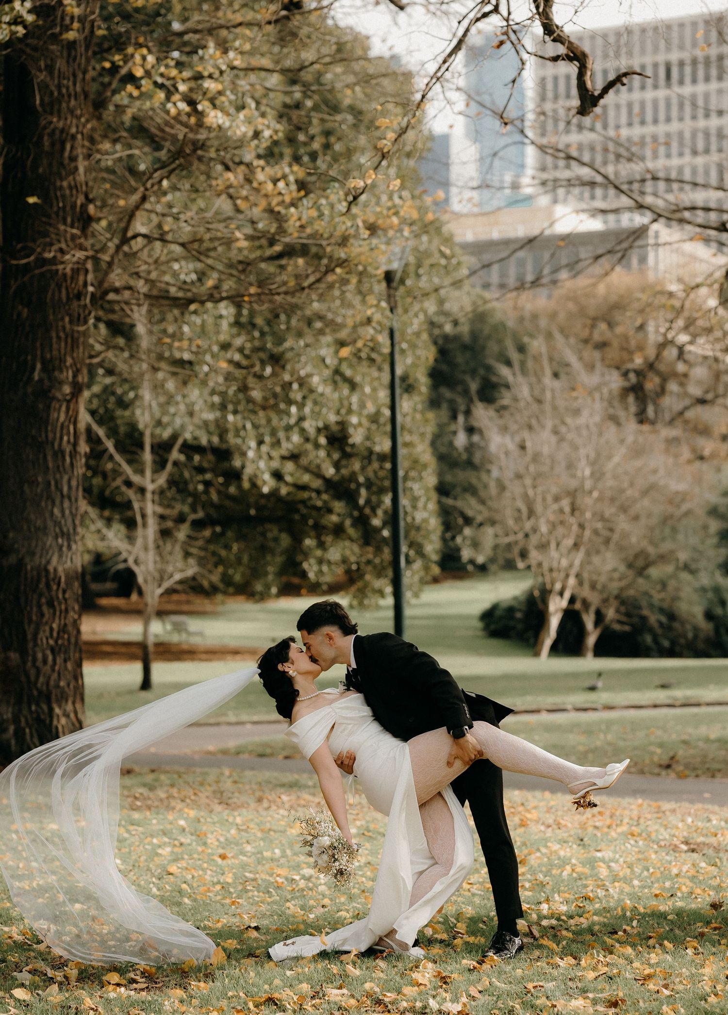 Bride and groom share a romantic dip kiss in an autumn park, with golden leaves and city skyline in the background.