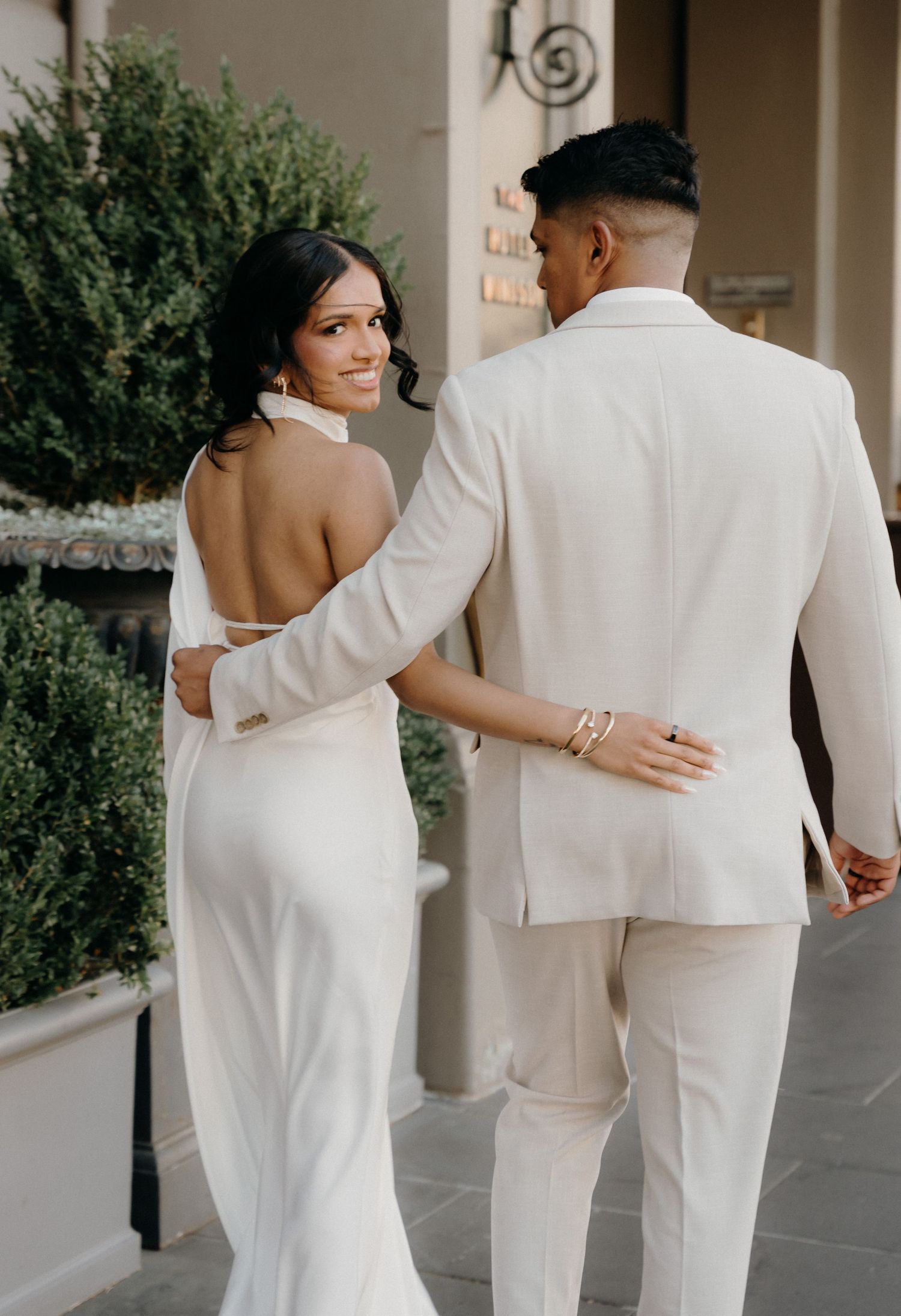 A couple in white wedding attire walking together and embracing in a series of candid photos along a covered walkway.