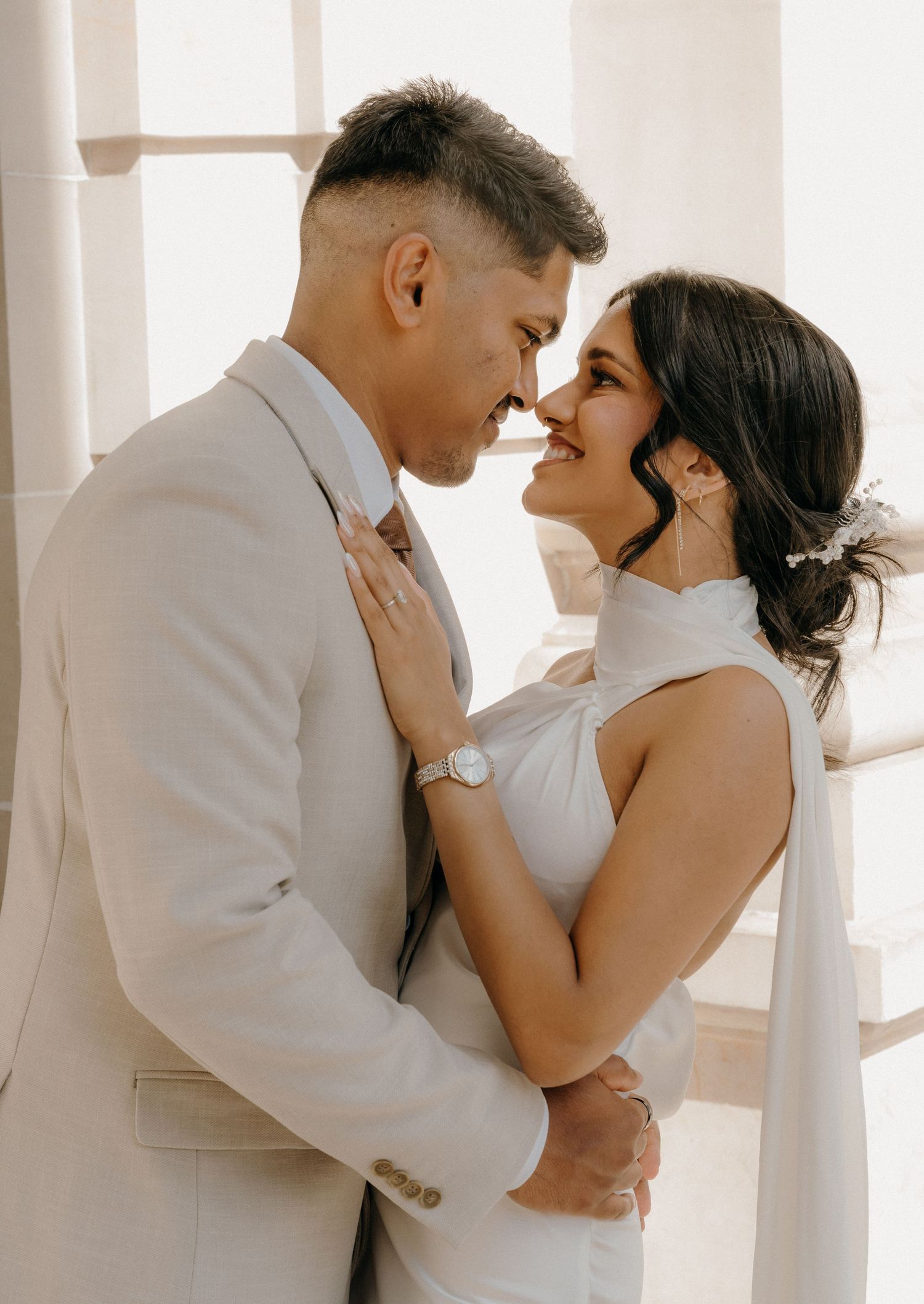 A romantic couple in white attire embraces in a bright sunlit room with elegant white columns.