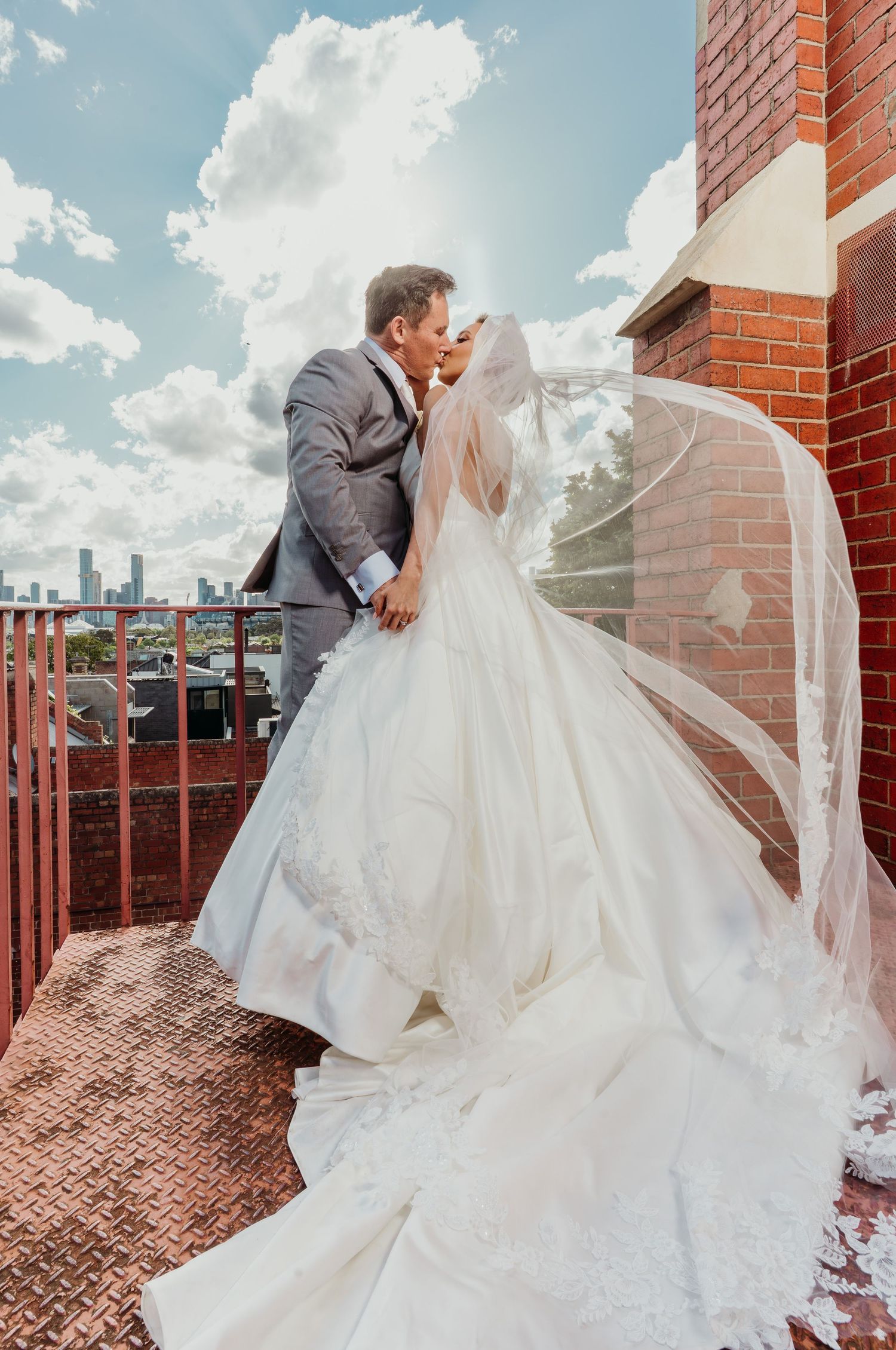 A bride and groom share a romantic moment on a rooftop terrace with a flowing veil and brick walls in the background.