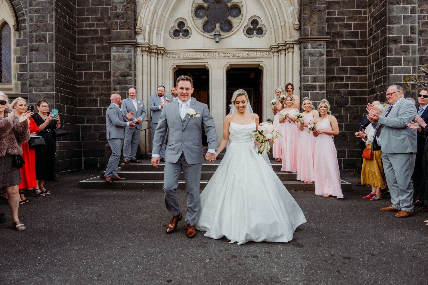 Wedding party poses in front of stone church entrance with guests watching from the sides.