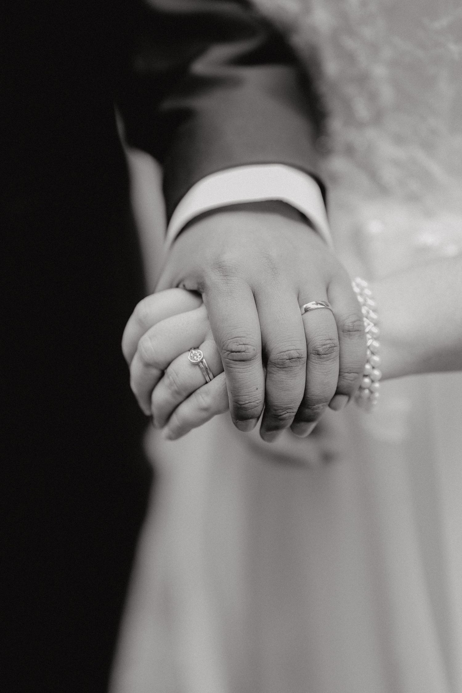 Black and white close-up photo of clasped hands wearing wedding rings and a pearl bracelet.