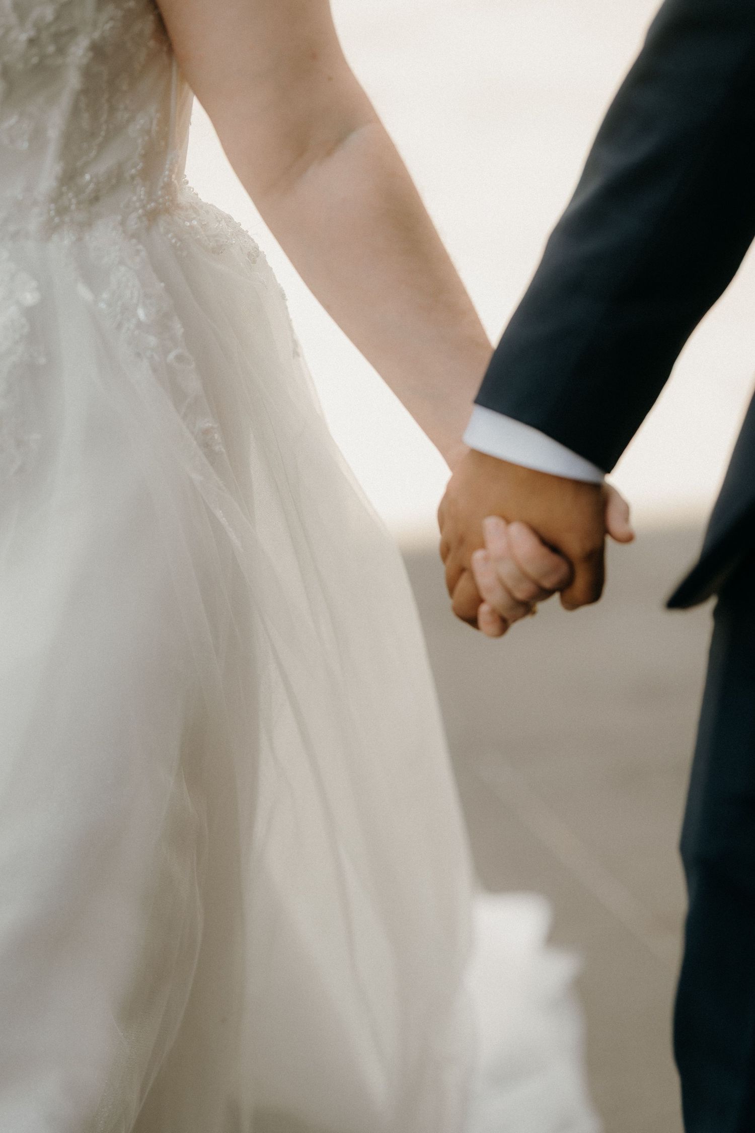 A bride and groom hold hands together in a soft, romantic wedding photo showing white dress and black suit details.