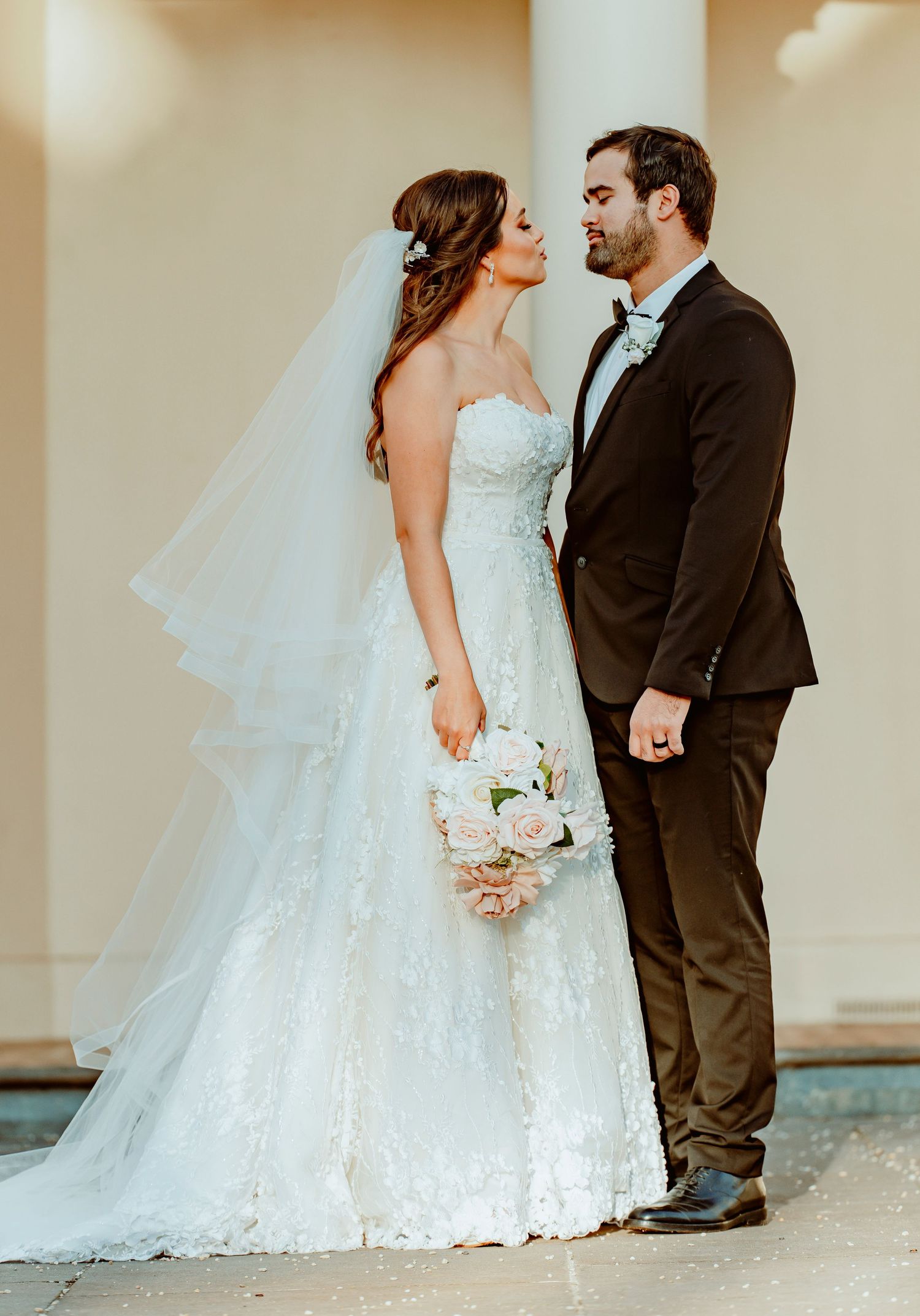A bride in a lace wedding gown holds a pink bouquet while sharing a romantic moment with groom in black tuxedo.