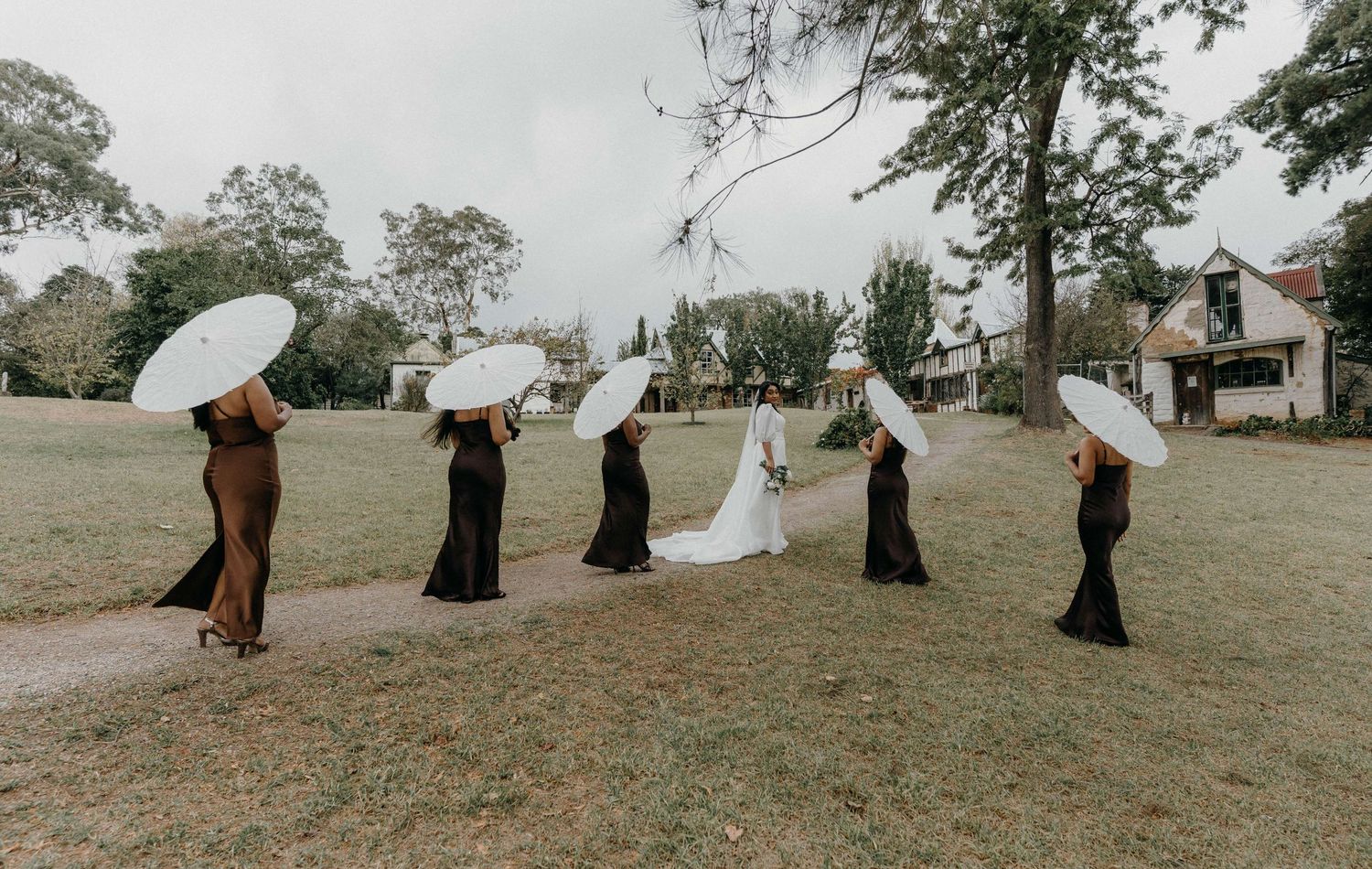 A wedding party stands in a line on a grassy lawn holding white umbrellas under an overcast sky.