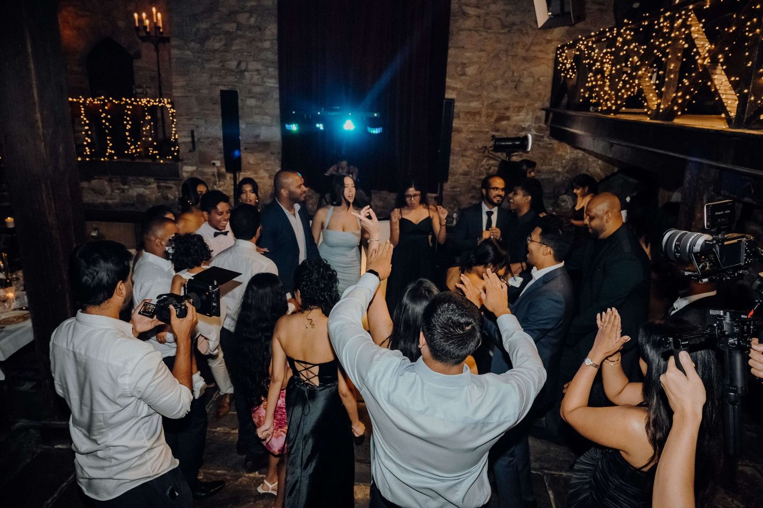 Wedding party dancing and celebrating on a dark dance floor with twinkling lights and photographers capturing the moment.