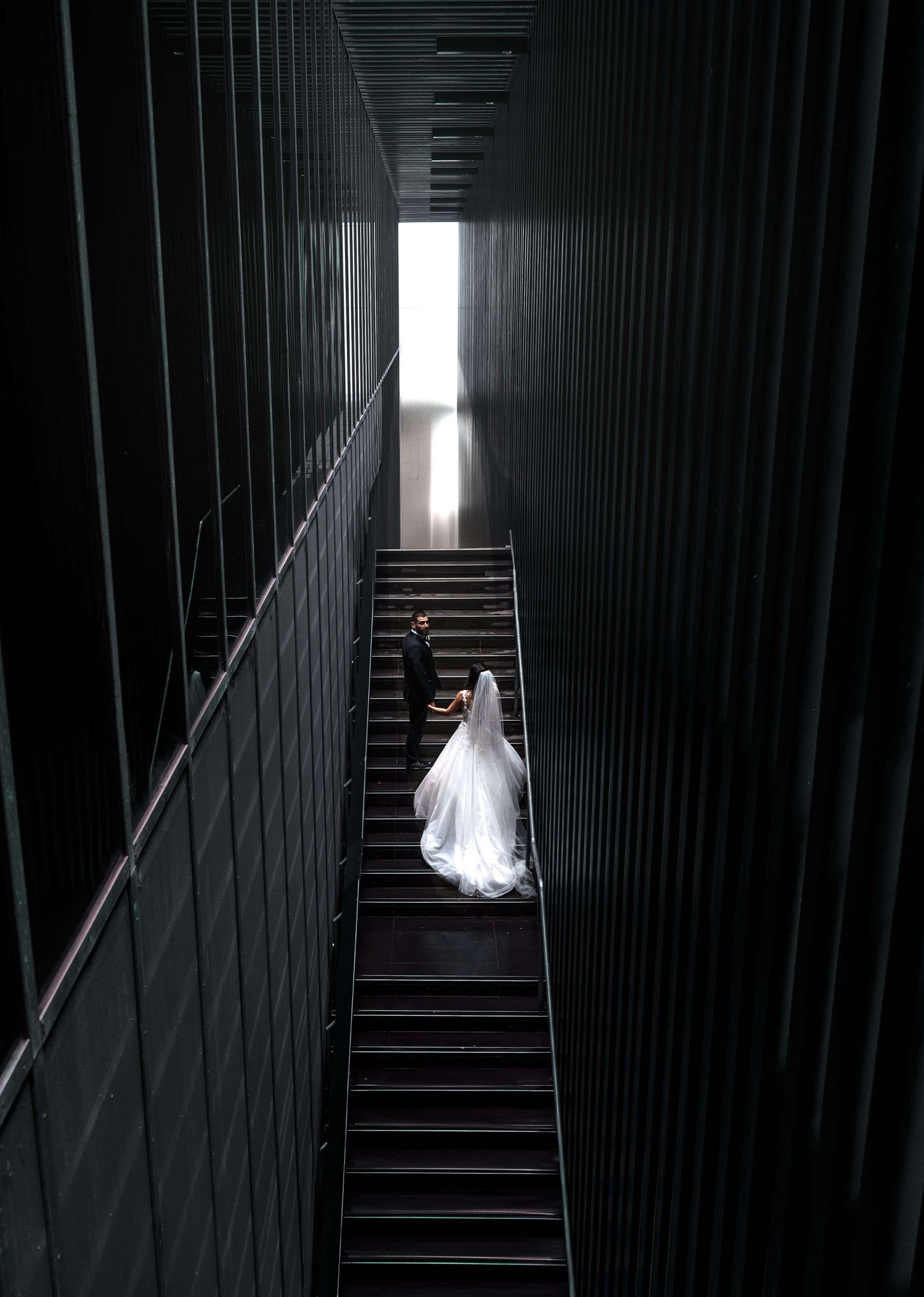 A person in a white wedding dress climbs dark industrial stairs in a dramatic architectural corridor with light at the end.