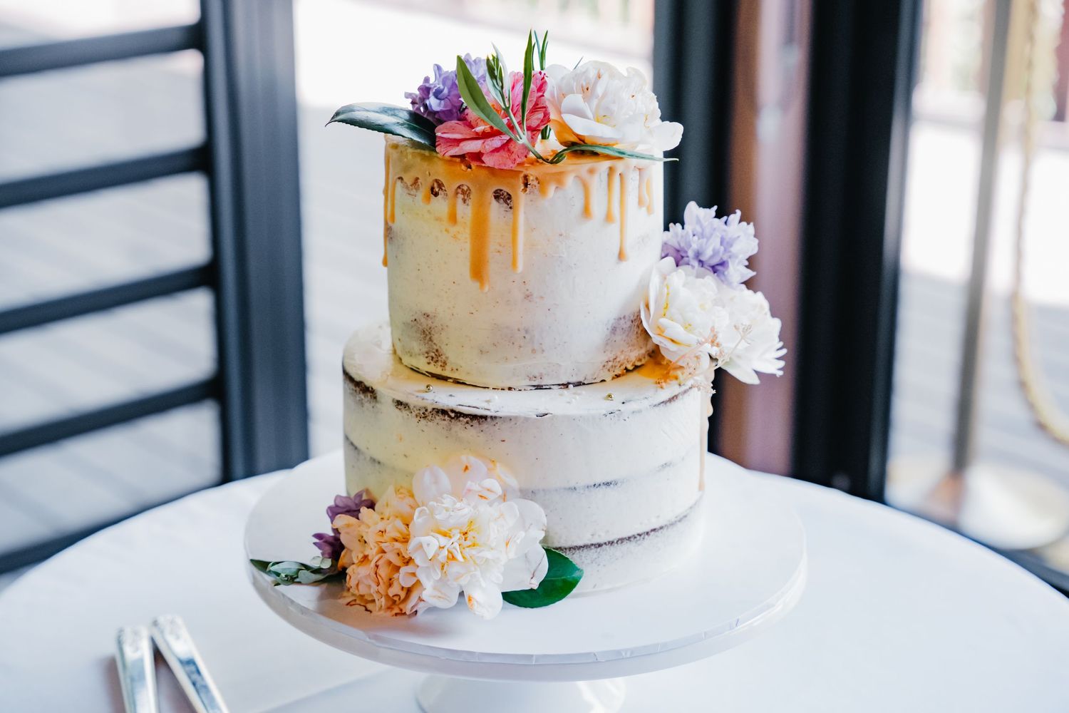 Elegant white wedding cake with fresh flowers and gold drip details displayed on a marble cake stand by a window.