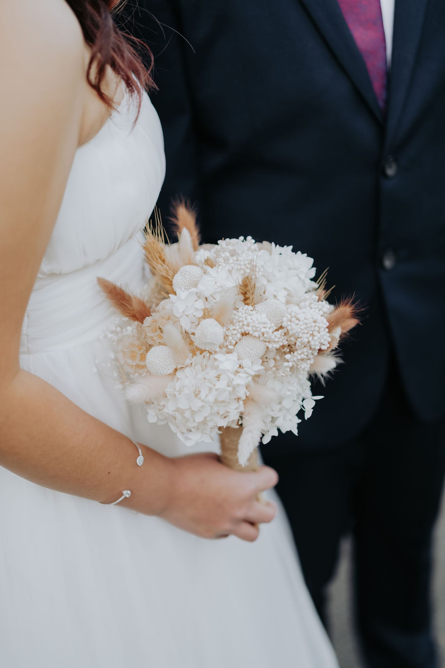 Detail shot of white bridal bouquet featuring dried flowers and delicate stems against a wedding dress background.