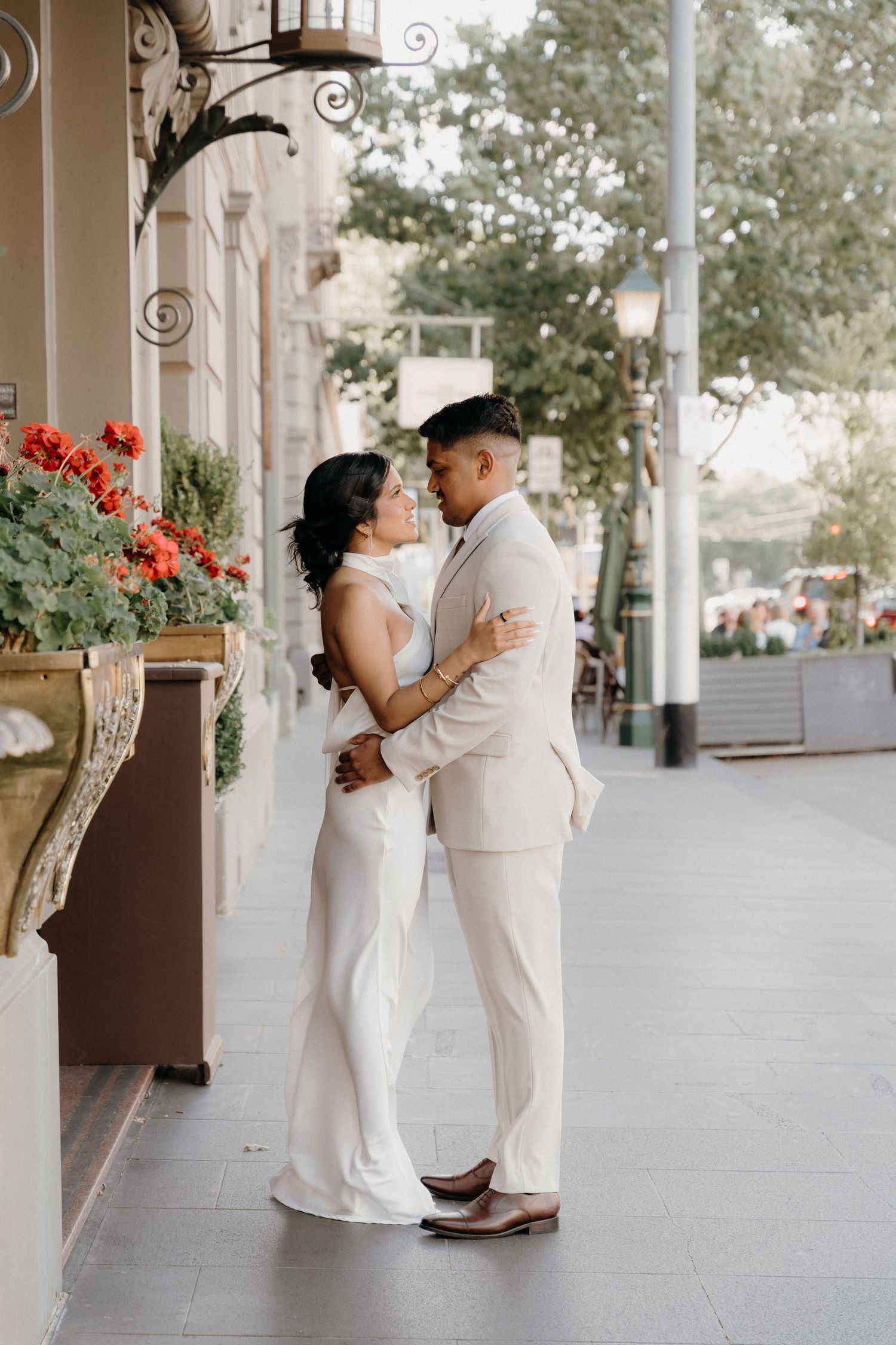 A romantic couple in matching white outfits embrace on a porch with red flowers and decorative architecture.
