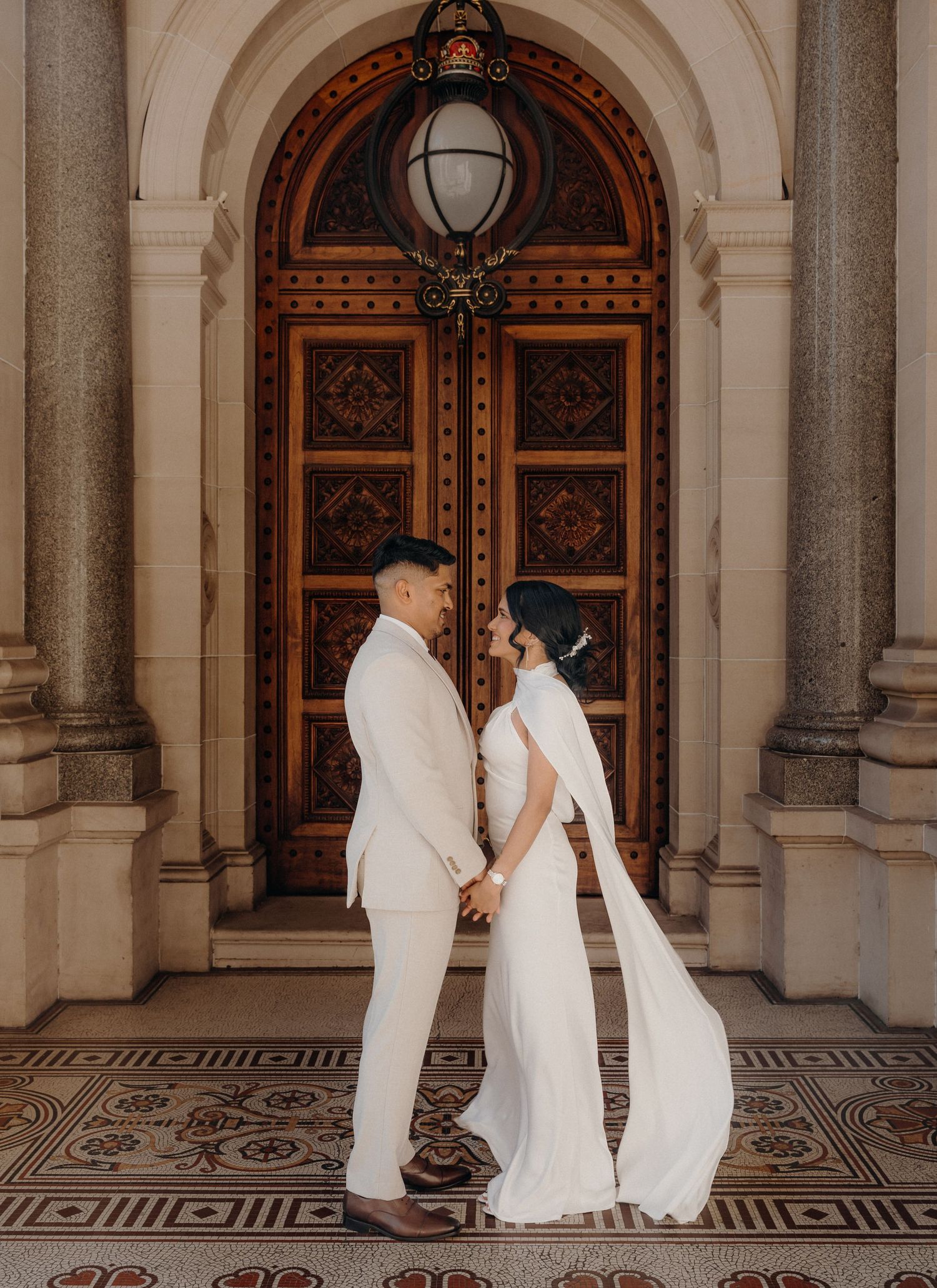 A couple in wedding attire shares romantic moments in an elegant archway corridor with ornate doors and tiled floors.