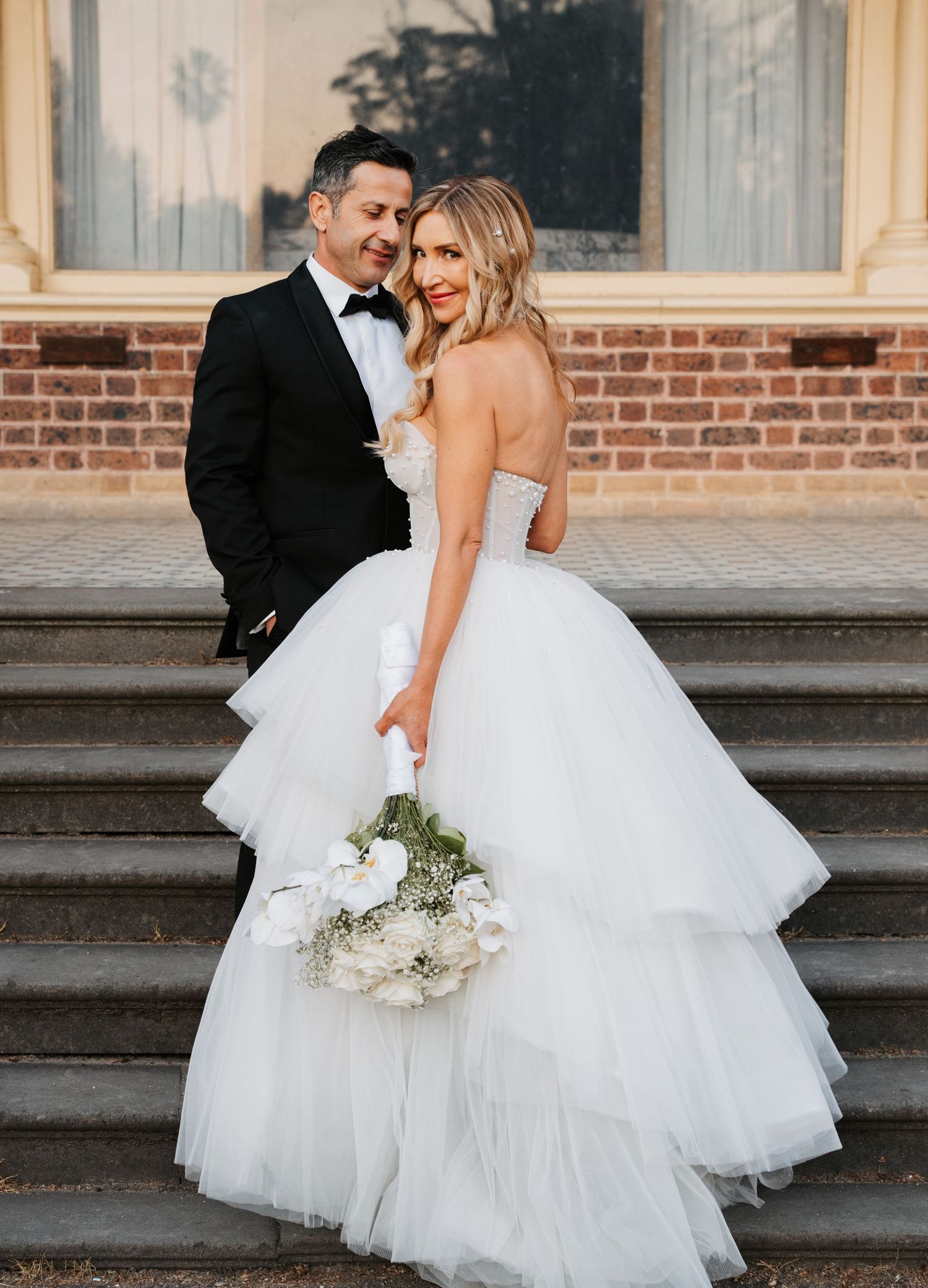 Romantic wedding couple embraces on stone steps with flowing white tulle gown and classic bouquet at sunset.