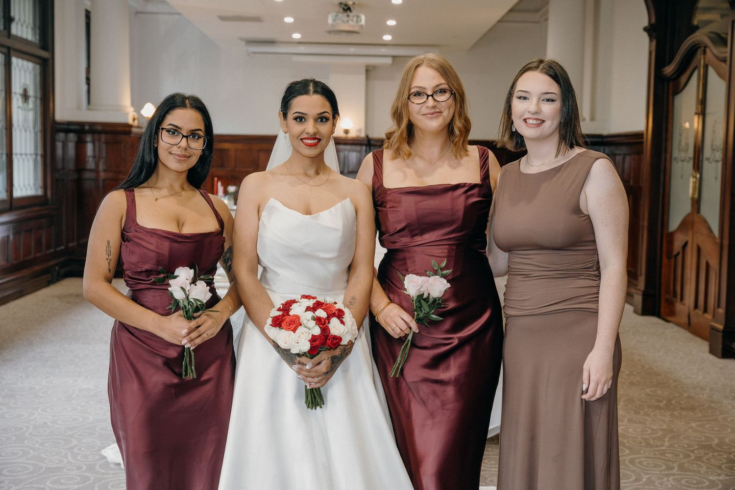 Bride in white gown poses with three bridesmaids in burgundy and taupe dresses holding bouquets in elegant venue.