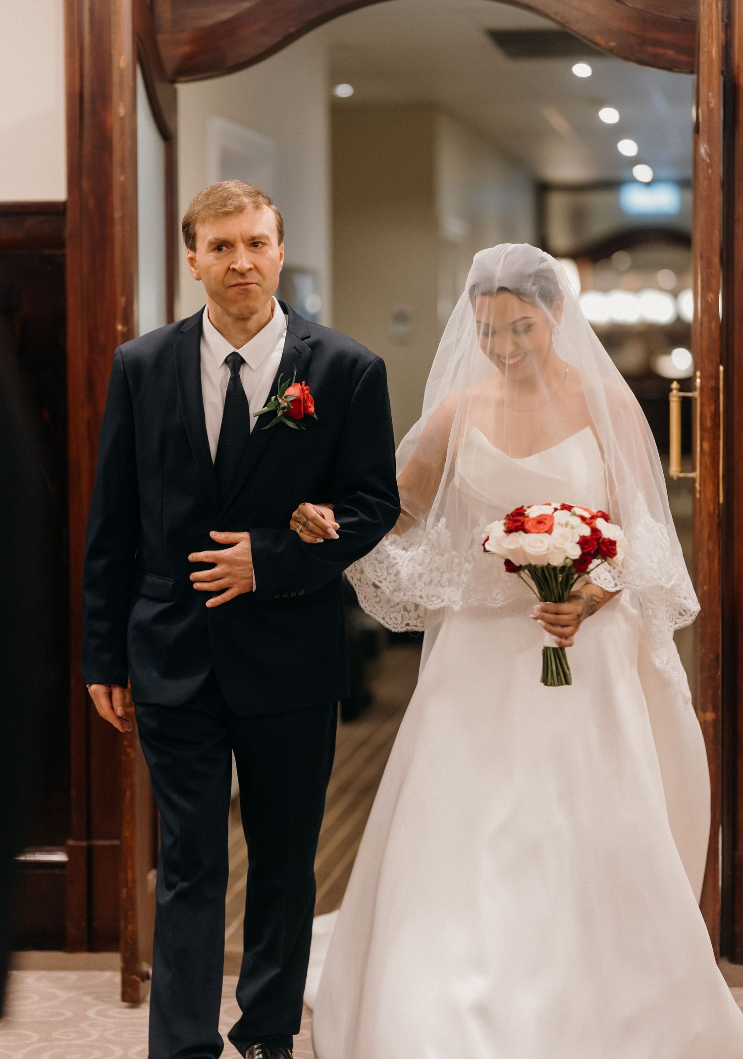 Bride in white gown and veil holding red bouquet walks down aisle with man in dark suit at wedding ceremony.