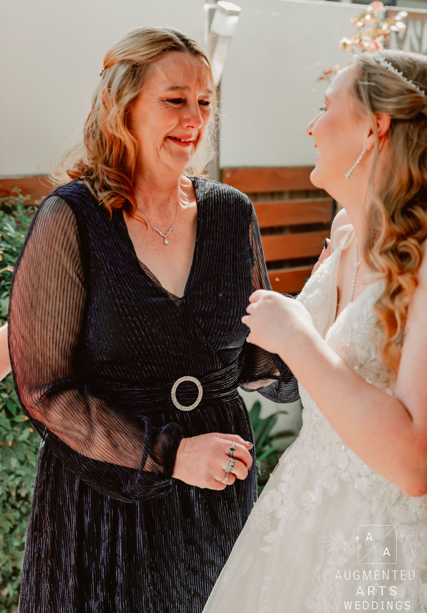 Candid moment between a woman in black dress and a bride in white gown sharing laughter during a wedding celebration.