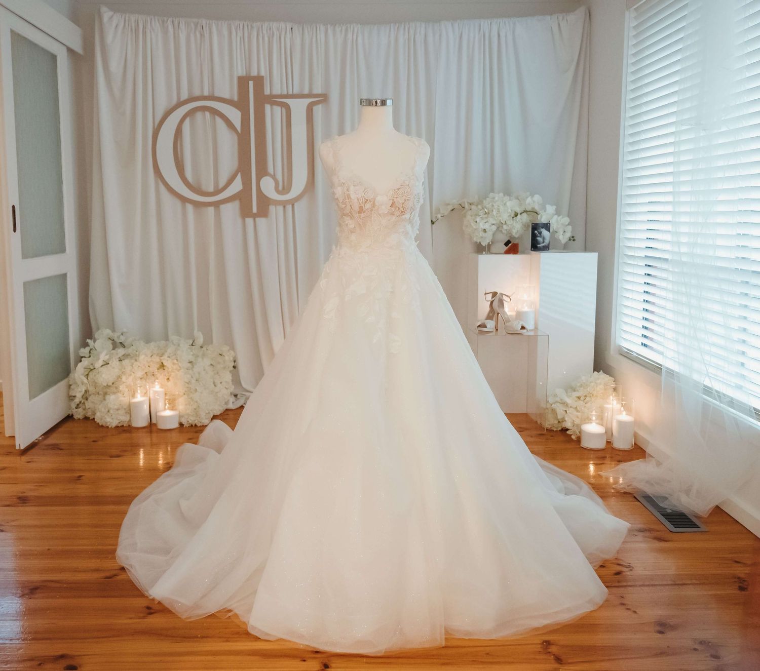 A collage of white wedding gowns with full tulle skirts displayed on dress forms in a sunlit room with hardwood floors.