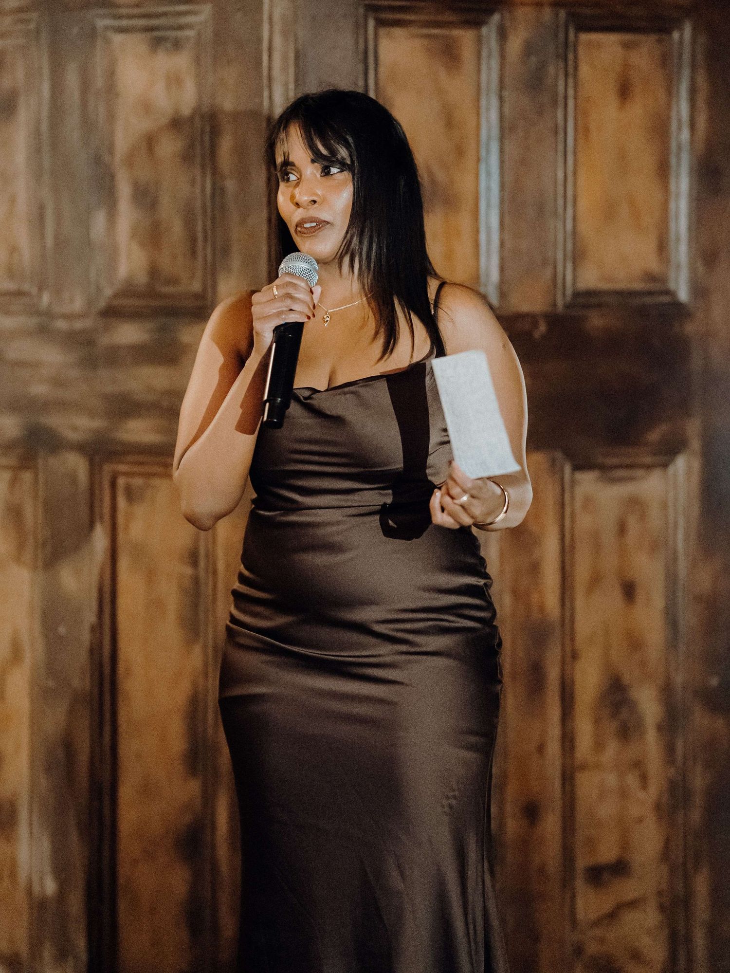 A woman in a brown satin dress holds a microphone against a wooden wall backdrop.