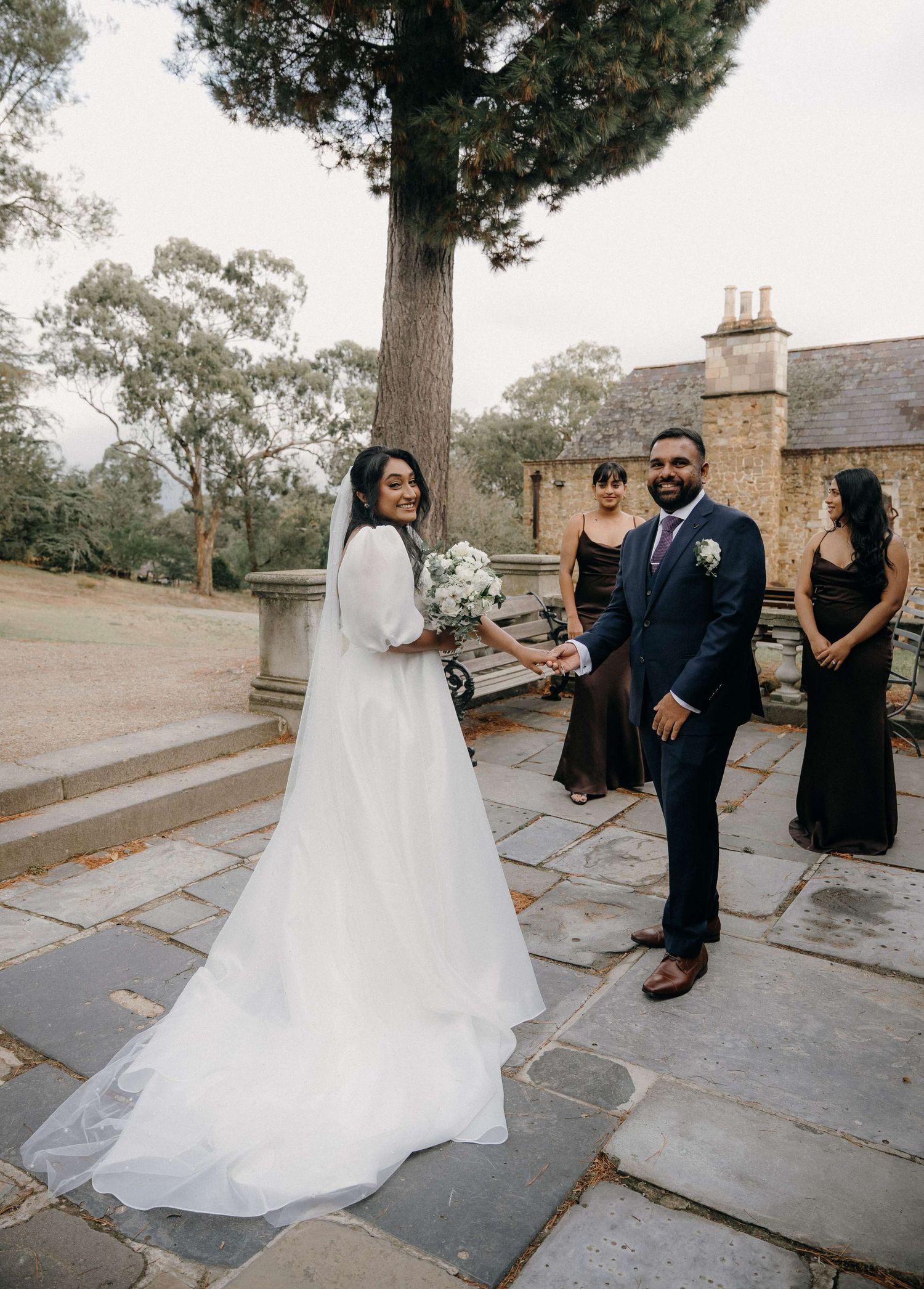 A flowing white wedding dress train sweeps across stone paving as figures in formal attire gather at a beautiful outdoor venue.