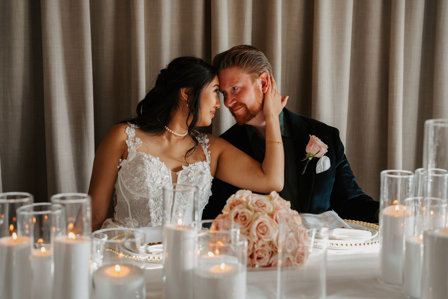 A romantic wedding couple shares an intimate moment at their candlelit reception table decorated with blush roses.