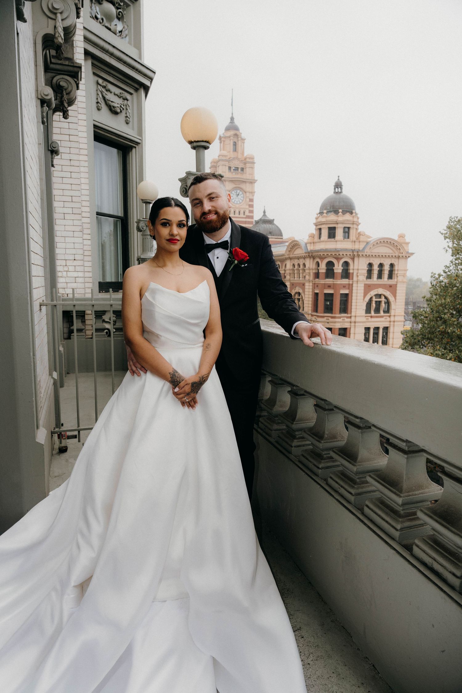 Bride in white ballgown and groom in black tuxedo pose on ornate balcony with Flinders Street Station backdrop.