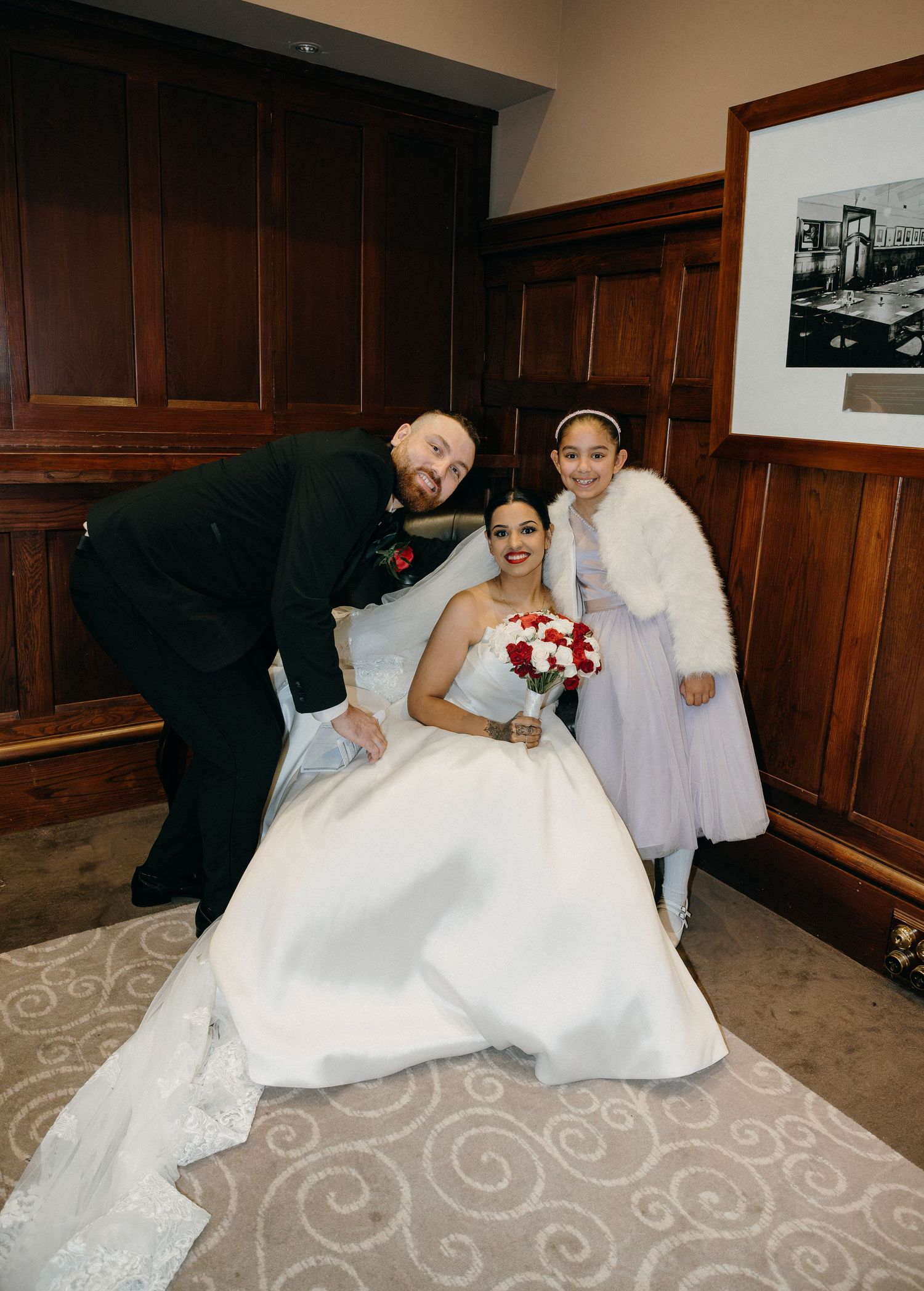 Bride in white ball gown holds red bouquet posing with groom and young flower girl in white dress indoors.