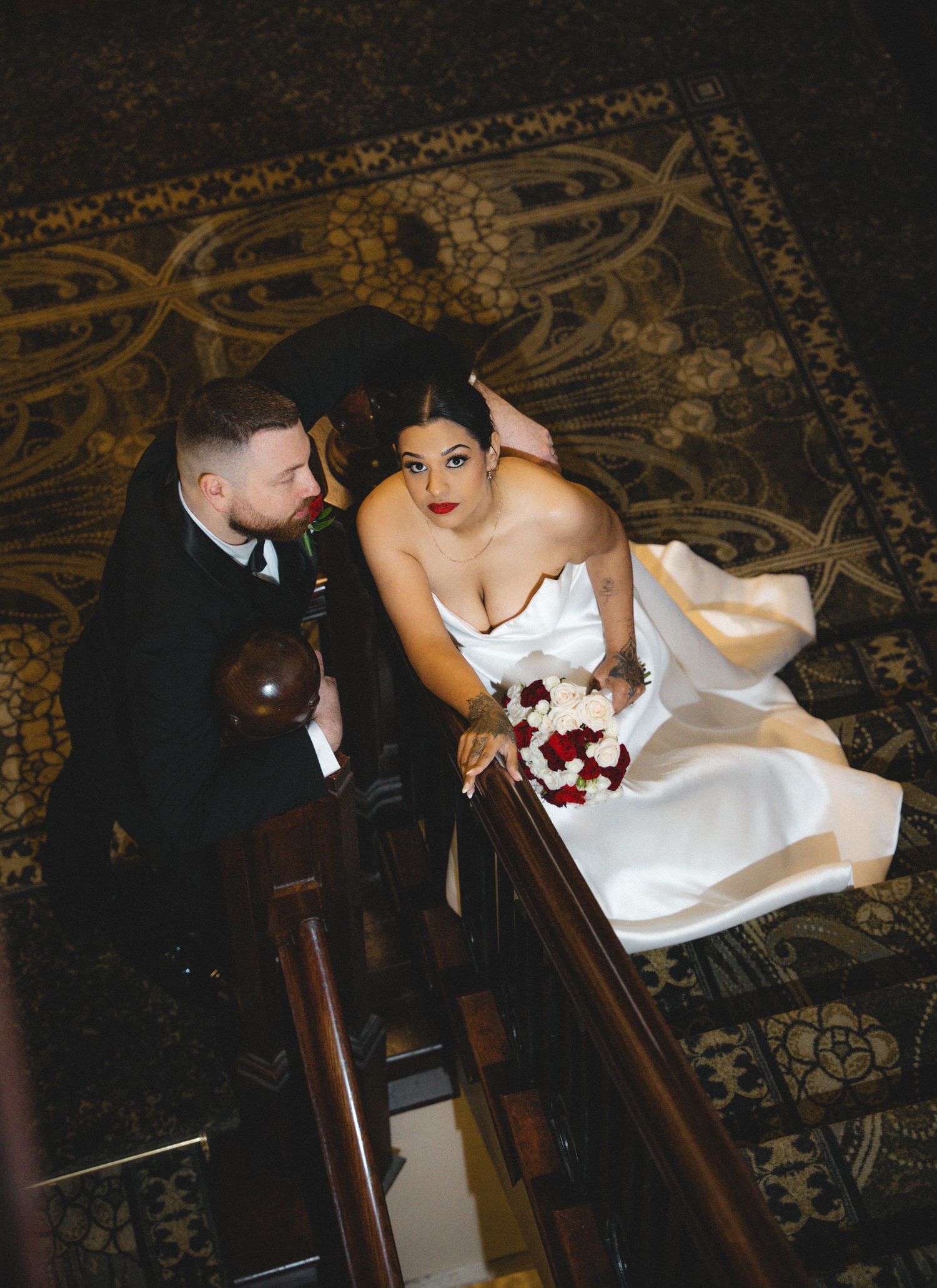 Bride in white gown holds red bouquet on ornate staircase beside groom in black suit, shot from above.