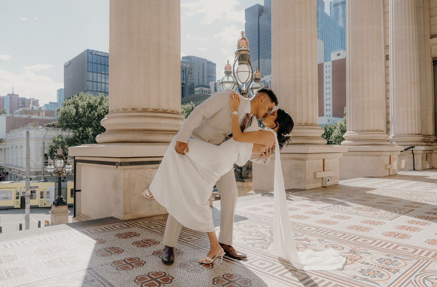 A person in white dress twirls gracefully through a grand marble colonnade with city buildings visible in the background.