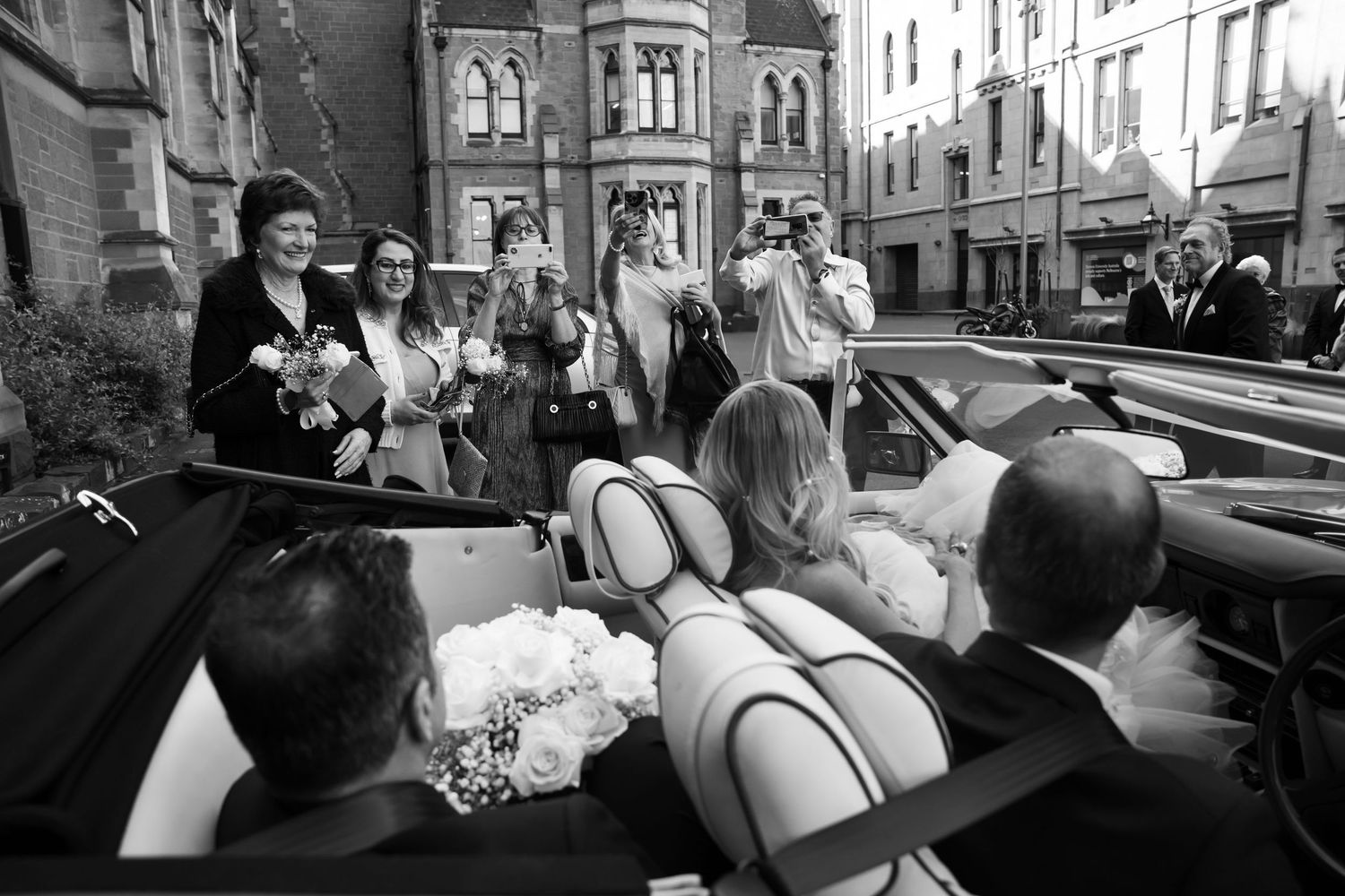 A wedding party celebrates in a vintage convertible car on a city street in black and white photography.