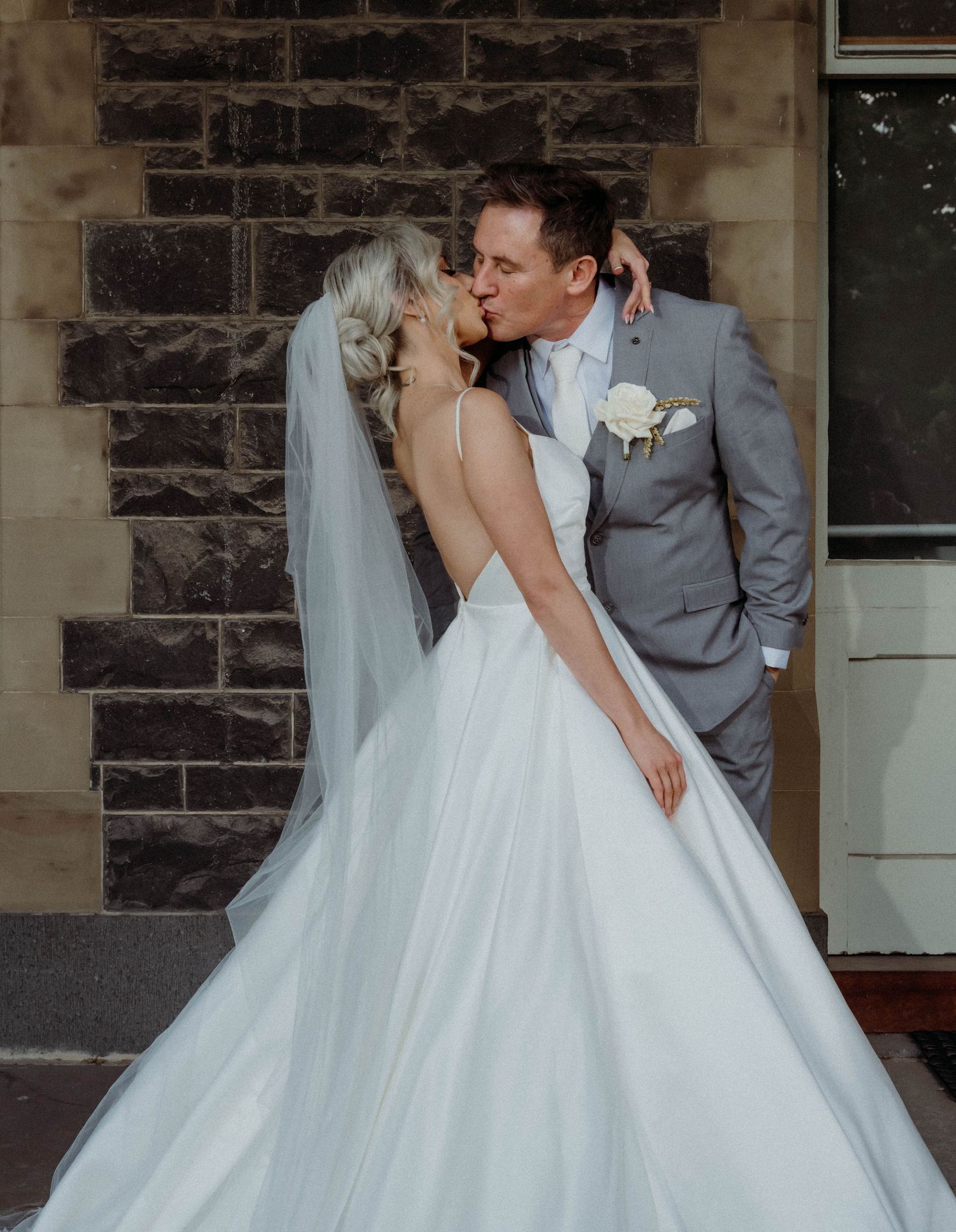 A romantic wedding couple shares a tender kiss in front of a stone building exterior.