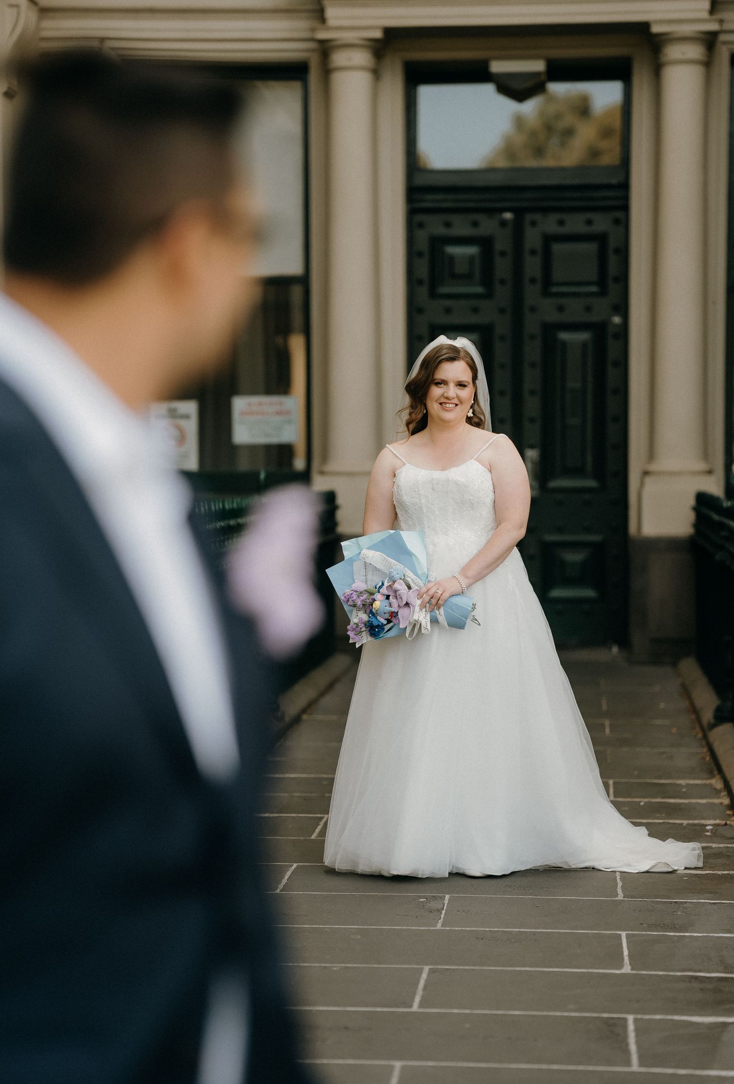 A bride in a white wedding gown holds a blue bouquet while standing in front of a grand stone building entrance.