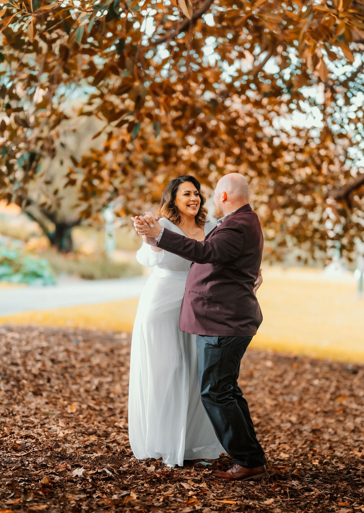 Couple dancing together in autumn leaves under trees with warm golden sunlight filtering through branches.