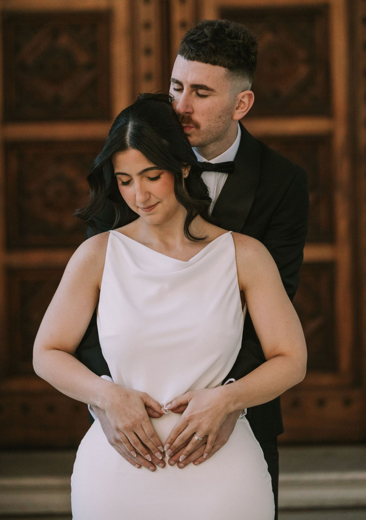 Bride in white satin gown embraced by groom in black tuxedo, posing tenderly before ornate wooden doors.