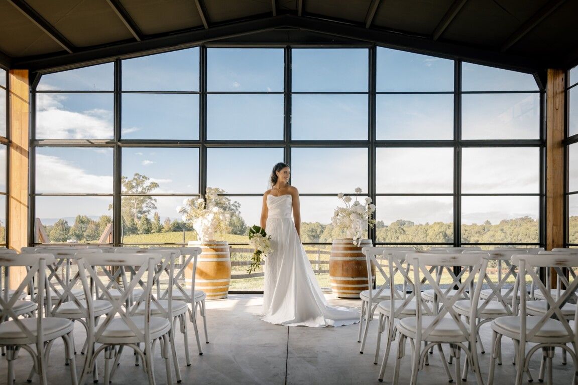 Bride in white gown stands between wine barrels in elegant venue with large windows and white chairs.